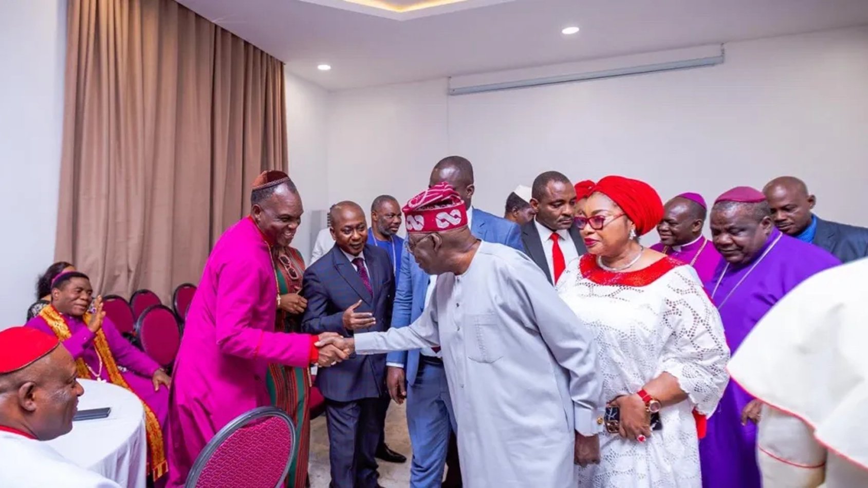 L'image montre un groupe de personnes rassemblées dans une salle, probablement lors d'un événement formel. Au centre, un homme en tenue traditionnelle serre la main d'un autre homme, qui porte une chemise claire. Les autres participants, habillés de manière variée, se tiennent autour d'eux. Certaines femmes portent des vêtements colorés et des accessoires élégants. L'ambiance semble conviviale et respectueuse, avec des sourires et des gestes amicaux. Les murs de la salle sont de couleur claire, et il y a des chaises rouges disposées autour de tables.