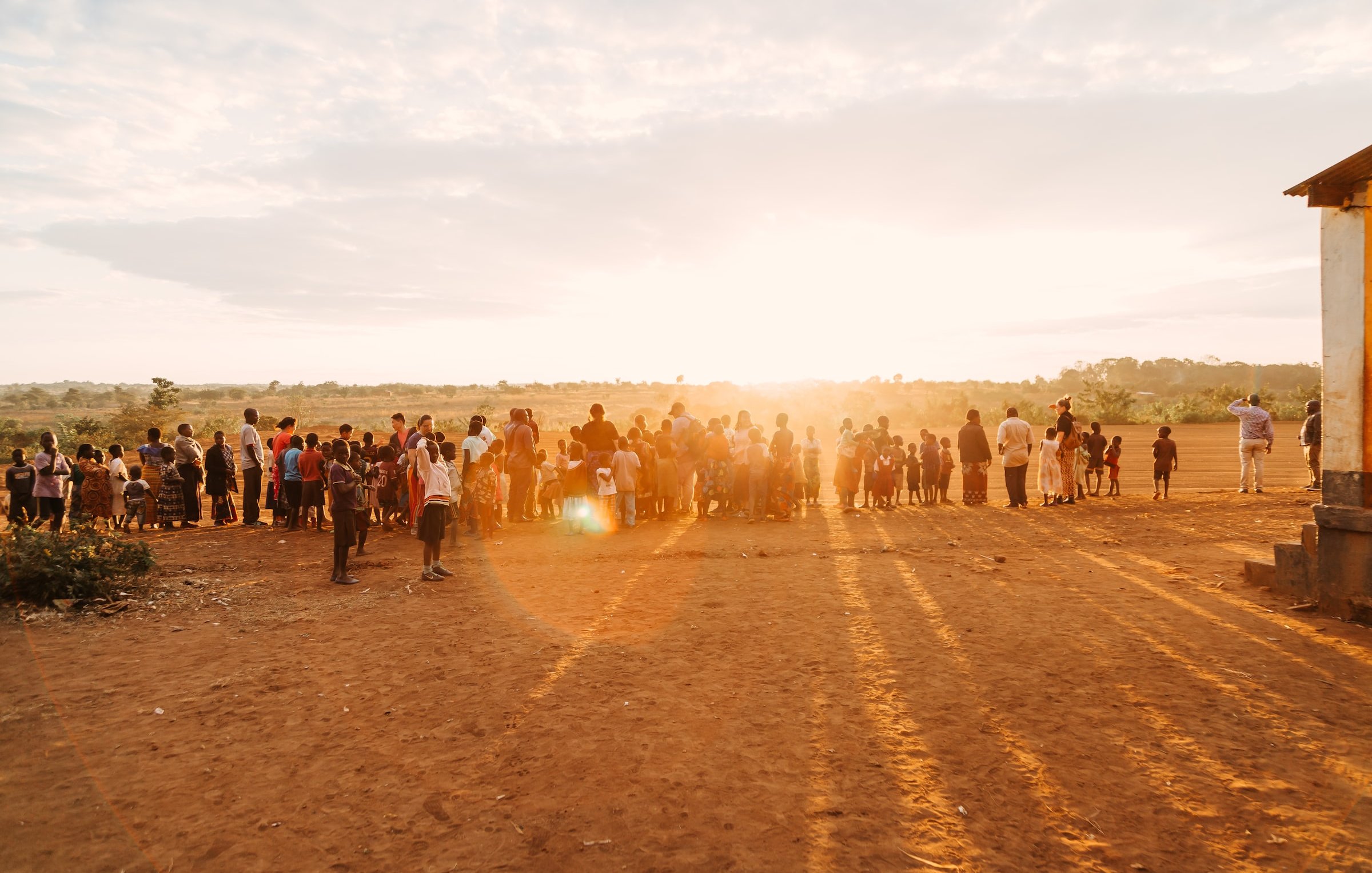 L'image présente une scène de rassemblement en plein air, au coucher du soleil. Au premier plan, un groupe nombreux de personnes se tient sur un terrain poussiéreux, probablement un village ou une communauté rurale. Les silhouettes des adultes et des enfants se distinguent distinctement alors qu'ils font face à l'horizon où le soleil descend, projetant de longues ombres sur le sol. L'arrière-plan est composé de plantes et d'une vaste étendue de paysage, créant une atmosphère paisible et chaleureuse. Le ciel, teinté de couleurs douces, suggère une fin de journée tranquille.