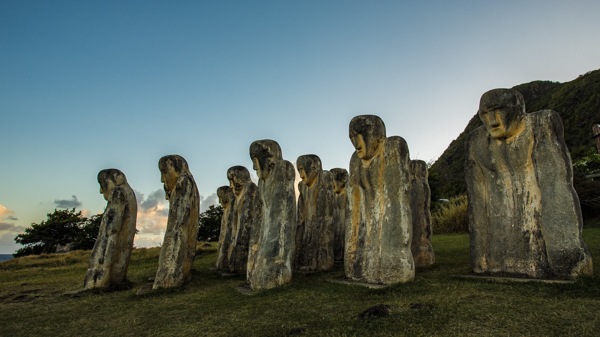 Cette image montre une série de statues de pierre, disposées en ligne sur un terrain herbeux. Les statues, représentant des figures humaines stylisées, se tiennent droites avec des traits faciaux peu définis et des postures légèrement inclinées, donnant une impression de profondeur et de mystère. En arrière-plan, on aperçoit des collines verdoyantes sous un ciel clair, où les nuances de bleu se fondent avec quelques nuages. La lumière du soleil, au coucher, illumine subtilement les statues, créant une atmosphère calme et contemplative, comme si ces figures immenses veillaient sur le paysage.