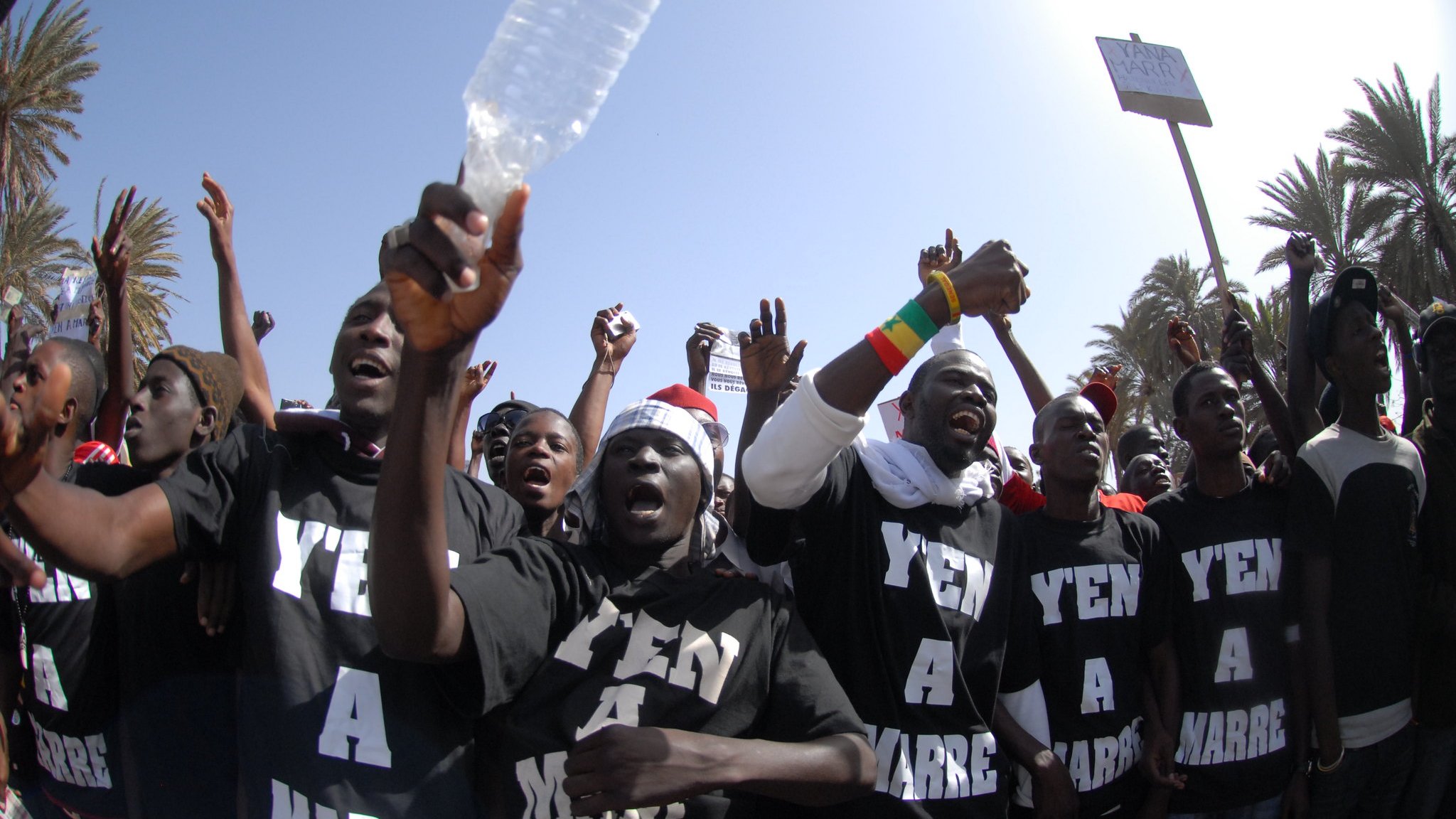 L'image montre un groupe de personnes rassemblées pour exprimer leur mécontentement, probablement lors d'une manifestation. Les membres du groupe portent des t-shirts noirs avec les mots "Y'en a marre" inscrits en lettres blanches, ce qui signifie qu'ils en ont assez d'une situation particulière. On peut les voir lever les bras, certains brandissant des bouteilles en plastique, et afficher des visages déterminés. En arrière-plan, on aperçoit des palmiers et un ciel bleu, indiquant une ambiance ensoleillée. L'énergie de la scène est palpable, avec des cris et une forte intensité émotionnelle.