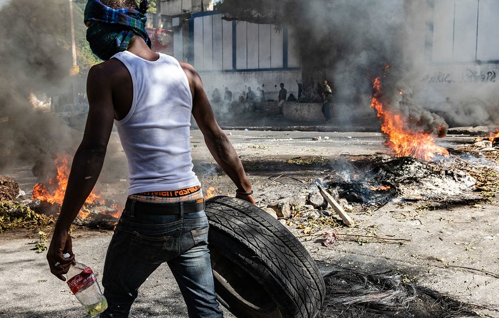 L'image montre une scène intense de protestation. Un homme, portant un débardeur blanc et un bandana sur le visage pour se protéger, est vu en arrière-plan. Il transporte un pneu que semble-t-il il va utiliser pour le jeter dans un feu. Autour de lui, on aperçoit de la fumée épaisse s'élevant dans l'air, résultant de plusieurs feux allumés sur la route. Le sol est jonché de débris, et l'atmosphère est chargée de tension, évoquant un sentiment de chaos et de lutte. Les flammes crépitent et laissent émaner une lumière orange. En arrière-plan, on peut distinguer des silhouettes de personnes, probablement d'autres manifestants.
