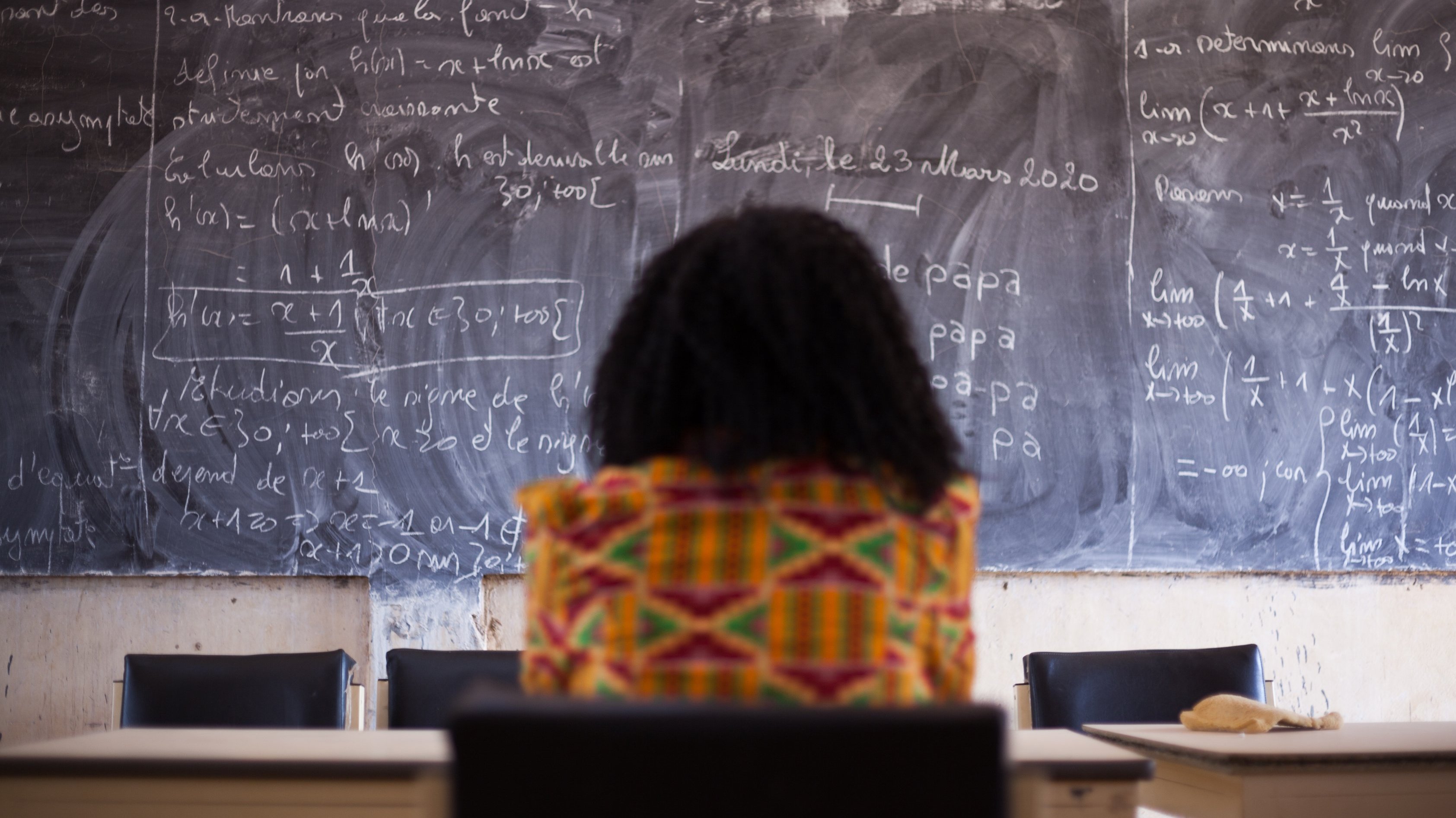 Dans cette image, on voit une personne assise, de dos, face à un grand tableau noir couvert d'écritures en craie blanche. Les formules mathématiques et les notes semblent complexes, suggérant un environnement académique ou d'apprentissage. La personne porte un vêtement coloré avec des motifs vibrants, ajoutant une touche de culture. L'ambiance est studieuse, et l'environnement évoque une salle de classe ou un espace de travail. Le contraste entre les couleurs vives du vêtement et la sombre surface du tableau crée une composition visuelle intéressante.