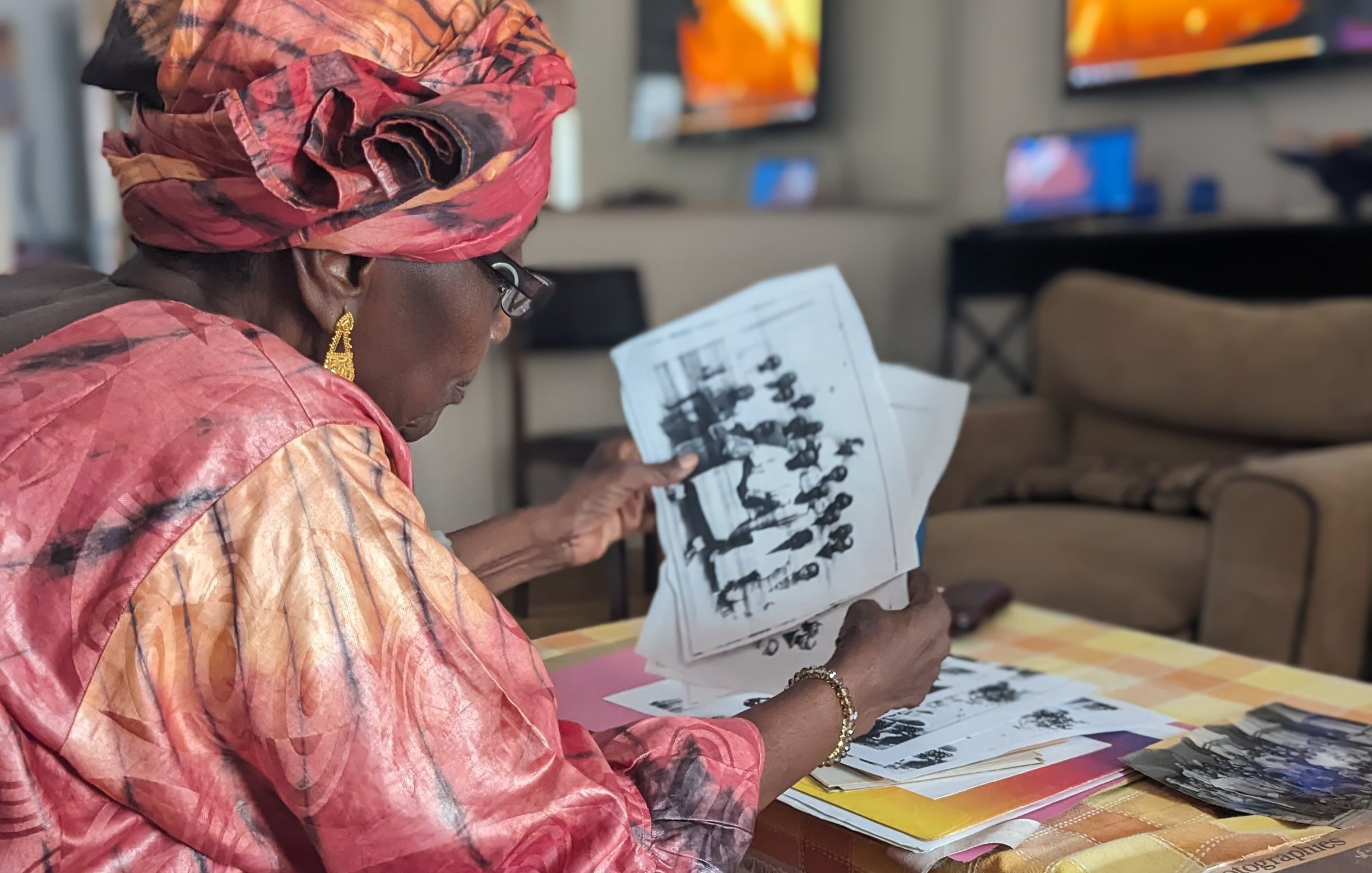 Dans cette image, une femme d'âge mûr, vêtue d'une magnifique robe rose et d'un foulard assorti, est assise sur un canapé dans un salon lumineux. Elle feuillette attentivement des photos en noir et blanc, toutes étalées devant elle sur une table basse recouverte d'une nappe à carreaux jaune et blanc. Le décor du salon est moderne, avec des télévisions visibles en arrière-plan, reflétant une ambiance chaleureuse et accueillante. La concentration de la femme témoigne de l'importance des souvenirs évoqués par ces images.