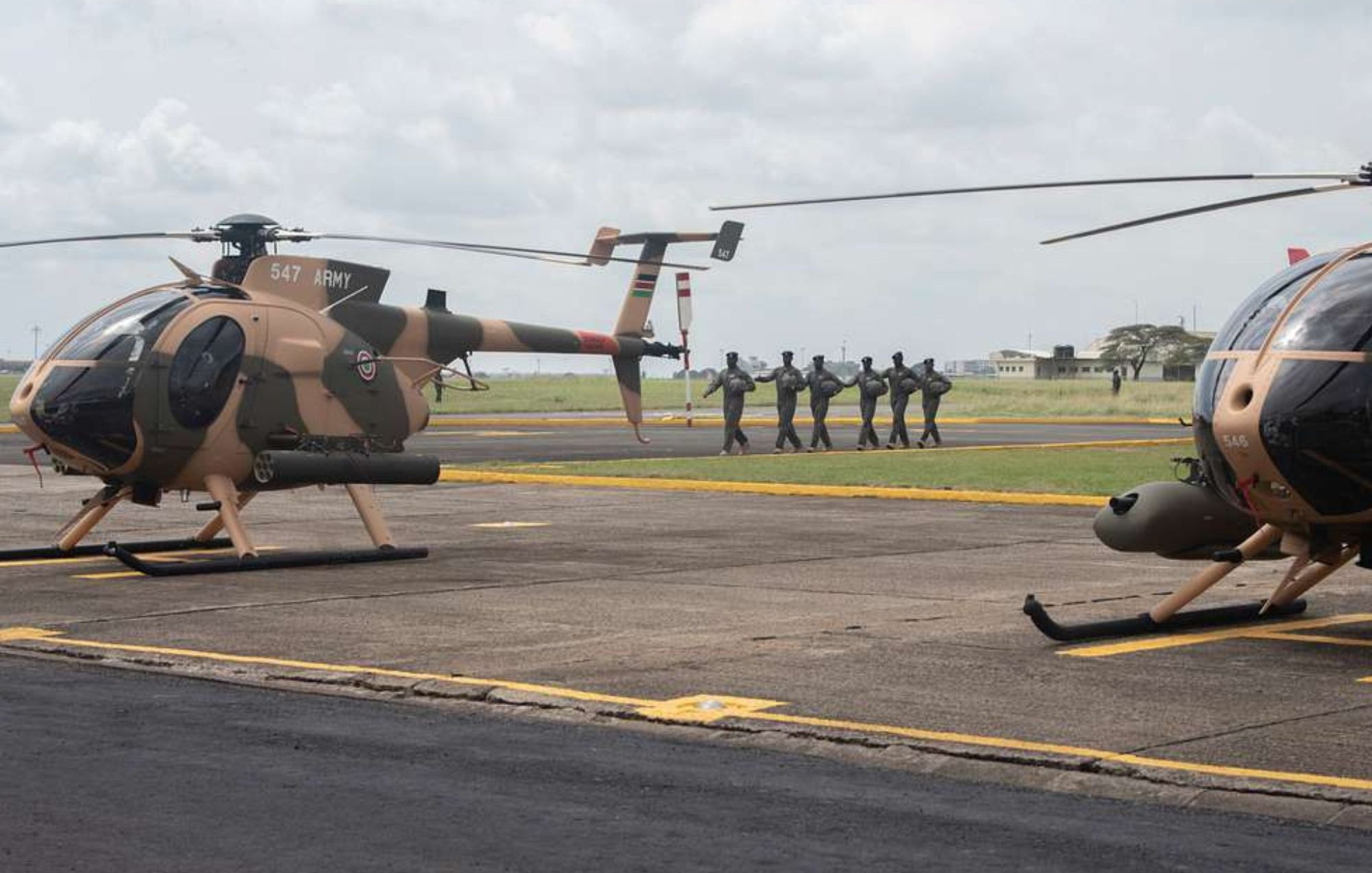 L'image montre une scène aérienne sur un terrain d'aviation. Deux hélicoptères, de couleur beige avec des motifs camouflage, sont stationnés sur le tarmac. À l'arrière-plan, un groupe de soldats en uniforme se dirige en formation vers un point de destination, créant une atmosphère dynamique et militaire. Le ciel est nuageux, ajoutant une ambiance dramatique à la scène. Le paysage autour est dégagé, avec des bâtiments épars visibles au loin.