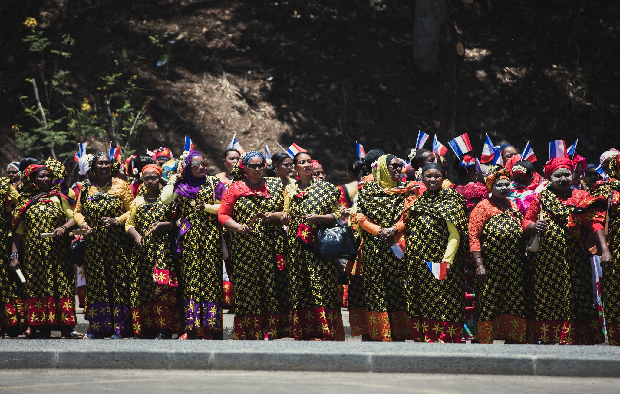 L'image montre un groupe de femmes réunies en ligne, vêtues de robes longues aux motifs floraux colorés. Chaque femme porte des accessoires traditionnels, comme des foulards vifs. Certaines tiennent des drapeaux aux couleurs bleu, blanc et rouge, symbolisant leur fierté nationale. L'arrière-plan est naturel, avec des arbres, et l'ambiance semble festive et engagée, suggérant une célébration ou un événement culturel. Les femmes affichent des expressions de joie et d'unité.