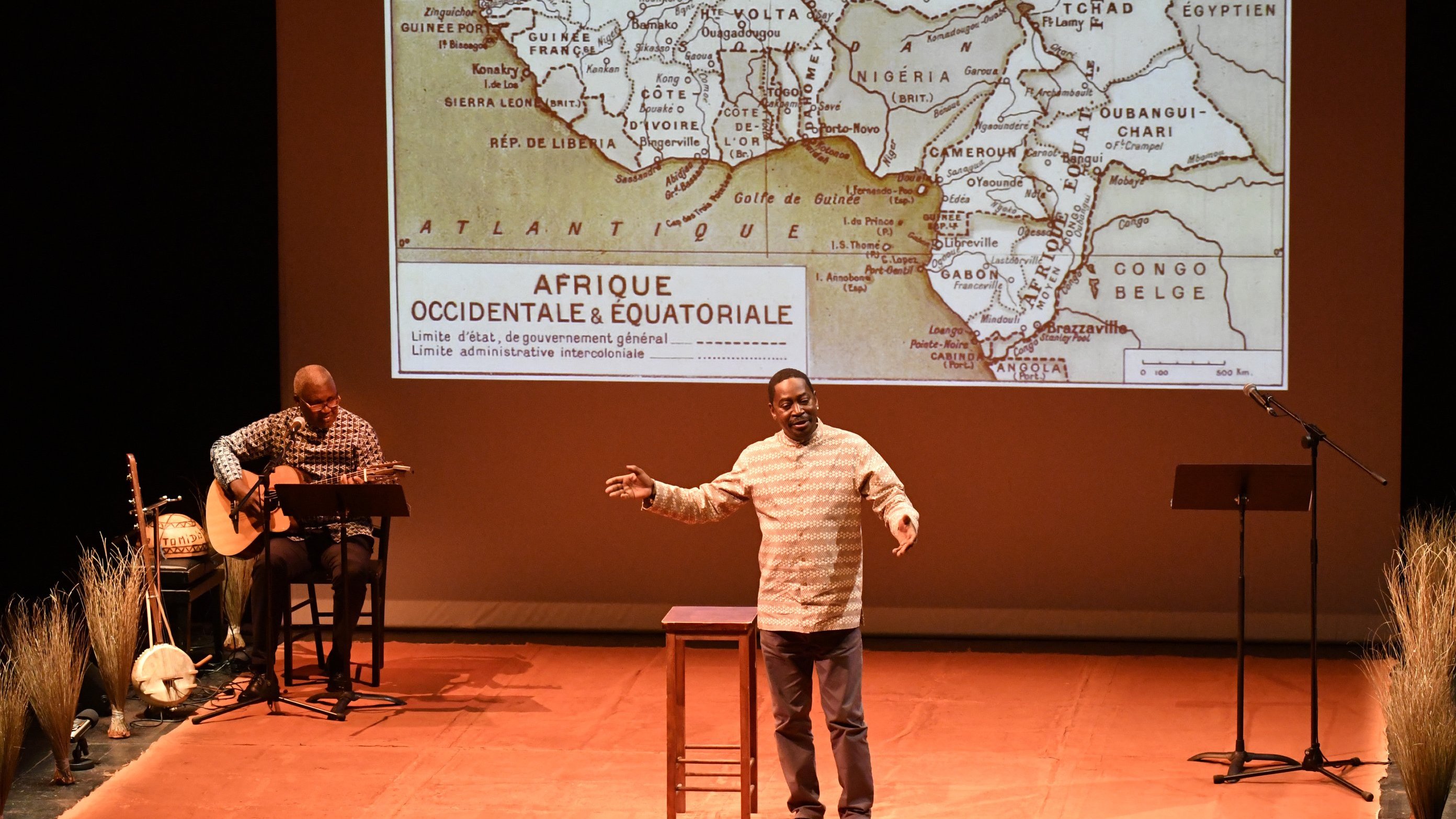 L'image représente un spectacle sur scène. Au premier plan, un homme s'exprime avec passion, les bras légèrement écartés pour accentuer son discours. Il porte une chemise claire et semble captivé par son sujet. À sa droite, un autre homme joue de la guitare assis sur un tabouret, ajoutant une ambiance musicale à la performance. En arrière-plan, un grand écran projette une carte ancienne de l'Afrique, spécifiquement d'Afrique occidentale et équatoriale, avec des contours de pays et des indications historiques. Le sol de la scène est décoré de brins de paille, ajoutant une touche naturelle à la mise en scène. L'éclairage doux met en valeur les acteurs et crée une atmosphère intimiste.