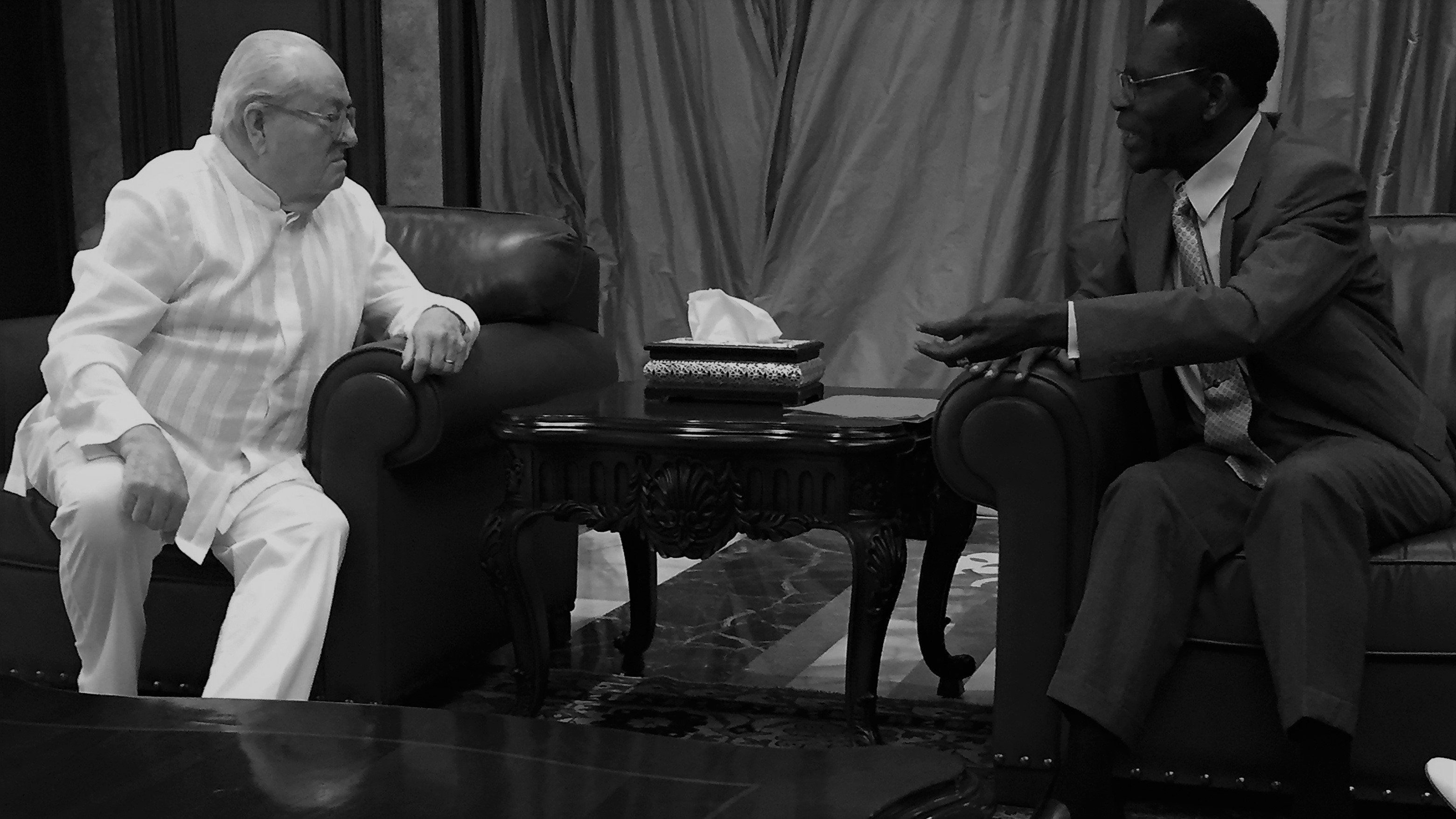 L'image représente une scène de discussion entre deux personnes assises l'une en face de l'autre dans un salon élégant. L'une des personnes porte un costume clair, tandis que l'autre est habillé d'un costume sombre. Ils sont assis sur des fauteuils en cuir, avec une table basse en bois ornée entre eux, où se trouve un objet décoratif. L'arrière-plan est composé de rideaux drapés et de murs en ton neutre, créant une atmosphère formelle et chaleureuse. L'éclairage est doux, ajoutant une sérénité à leur échange. On peut imaginer qu'ils discutent de manière engagée, leurs gestes accentuant l'intensité de la conversation.