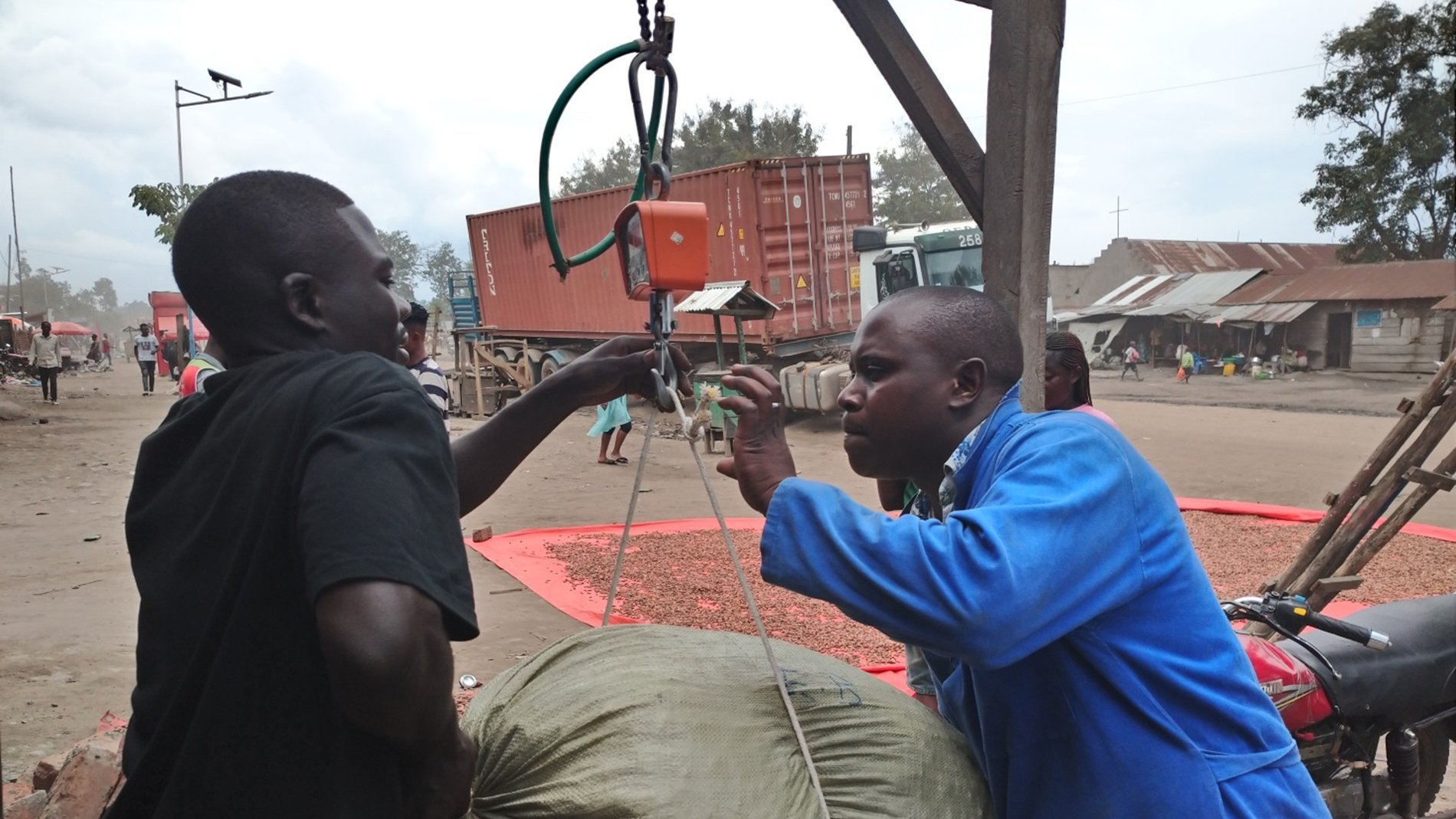 Dans cette image, deux hommes interagissent dans un environnement de marché. L'un des hommes, en tenue bleue, est en train de peser un gros sac à l'aide d'une balance suspendue. Le sac, de couleur beige et légèrement usé, semble lourd. L'autre homme, portant un t-shirt noir, aide à tenir le sac pendant qu'il est pesé. En arrière-plan, on peut apercevoir des conteneurs de transport et des structures typiques d'un marché, avec d'autres personnes qui se déplacent. L'atmosphère est animée, avec des sons de conversation et les bruits d'un marché en pleine activité.