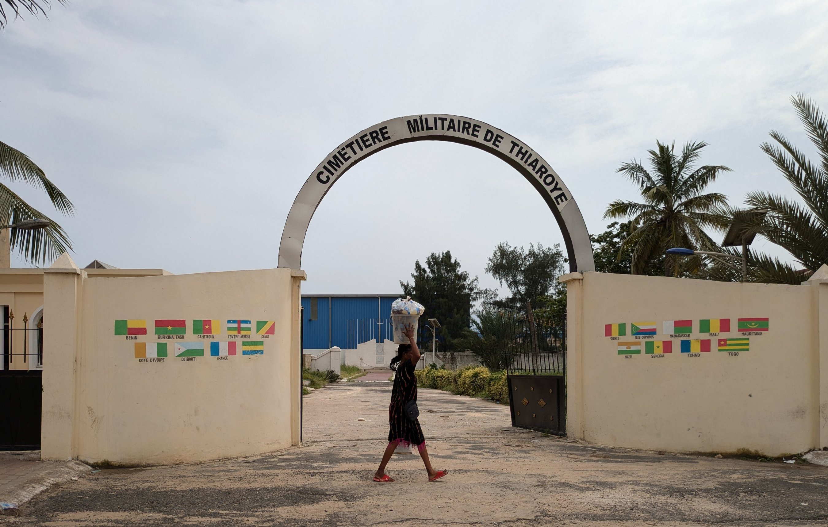 L'image montre l'entrée du cimetière militaire de Thiaroye. Un grand arc de portail est inscrit avec le nom "CIMETIÈRE MILITAIRE DE THIAROYE". En arrière-plan, on peut apercevoir quelques arbres, dont des palmiers, qui ajoutent une touche tropicale à la scène. Au premier plan, une personne marche, portant un grand récipient sur sa tête. Le sol est en béton, avec des traces de circulation. De part et d'autre de l'entrée, des murs affichent des drapeaux de différentes nations, apportant une sensation de diversité. L'ensemble dégage une ambiance calme et respectueuse.