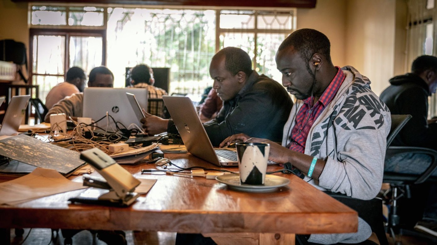 Dans cette image, on voit un espace de travail collaboratif. Plusieurs personnes sont assises autour d'une grande table en bois. Chaque personne utilise un ordinateur portable, principalement des MacBook. Il y a des câbles qui traînent sur la table, ajoutant à l'ambiance de travail intensif. À droite, un homme porte un sweat à capuche gris et écoute des écouteurs, concentré sur son écran. Sur la table, on aperçoit une tasse, peut-être de café, ainsi que quelques feuilles. Les murs sont clairs et de grandes fenêtres laissent entrer la lumière naturelle, créant une atmosphère chaleureuse et productive. En arrière-plan, d'autres personnes sont visibles, toutes absorbées dans leur travail.
