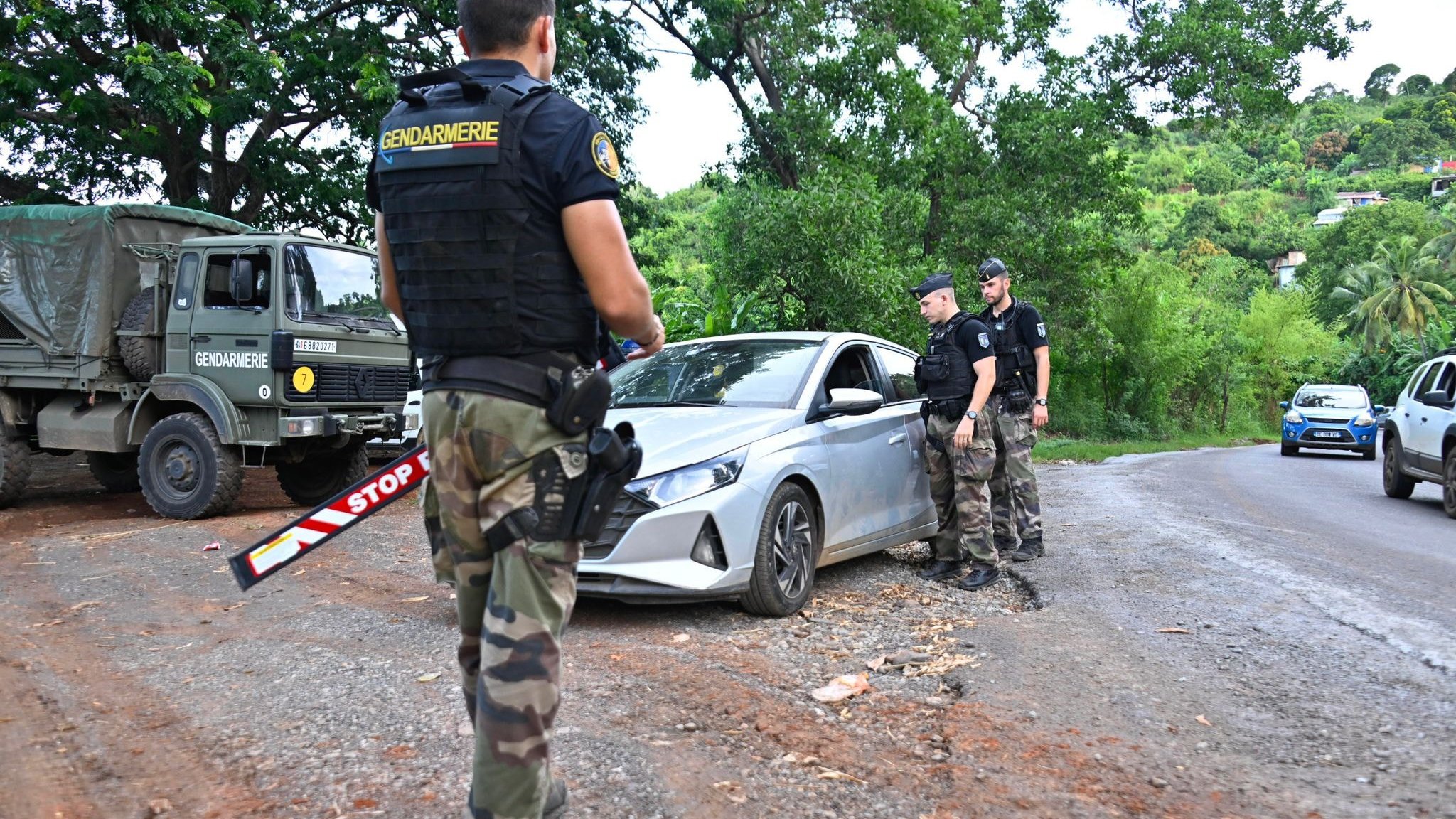 L'image montre un contrôle routier en plein air. Au premier plan, un gendarme en uniforme, portant un gilet par balles noir avec l'inscription "Gendarmerie", observe une voiture argentée qui est arrêtée sur le côté de la route. La voiture a un panneau "STOP" visible. Deux autres gendarmes, habillés de manière similaire, se tiennent près de la voiture, dialoguant probablement avec le conducteur. En arrière-plan, on aperçoit une camionnette militaire de la gendarmerie et des arbres verdoyants qui donnent une ambiance naturelle à la scène. Le sol est en terre, avec beaucoup de verdure autour, ce qui indique que le lieu est rural. L'atmosphère semble sérieuse, reflet du contrôle de sécurité en cours.