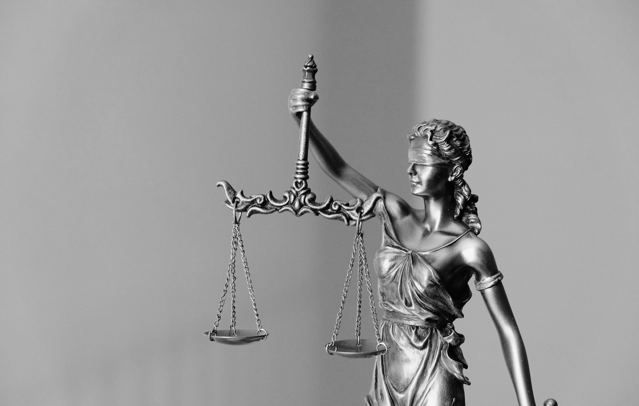 L'image représente une statue de la justice, souvent symbolisée par une femme. Elle est drapée dans une robe et tient une balance dans une main, symbolisant l'équilibre et l'équité. La statue est également dotée d'un bandeau sur les yeux, représentant l'impartialité dans le jugement. L'ensemble de la sculpture est réalisé dans des tons métalliques, et le fond est flou et neutre, mettant en valeur la figure centrale.