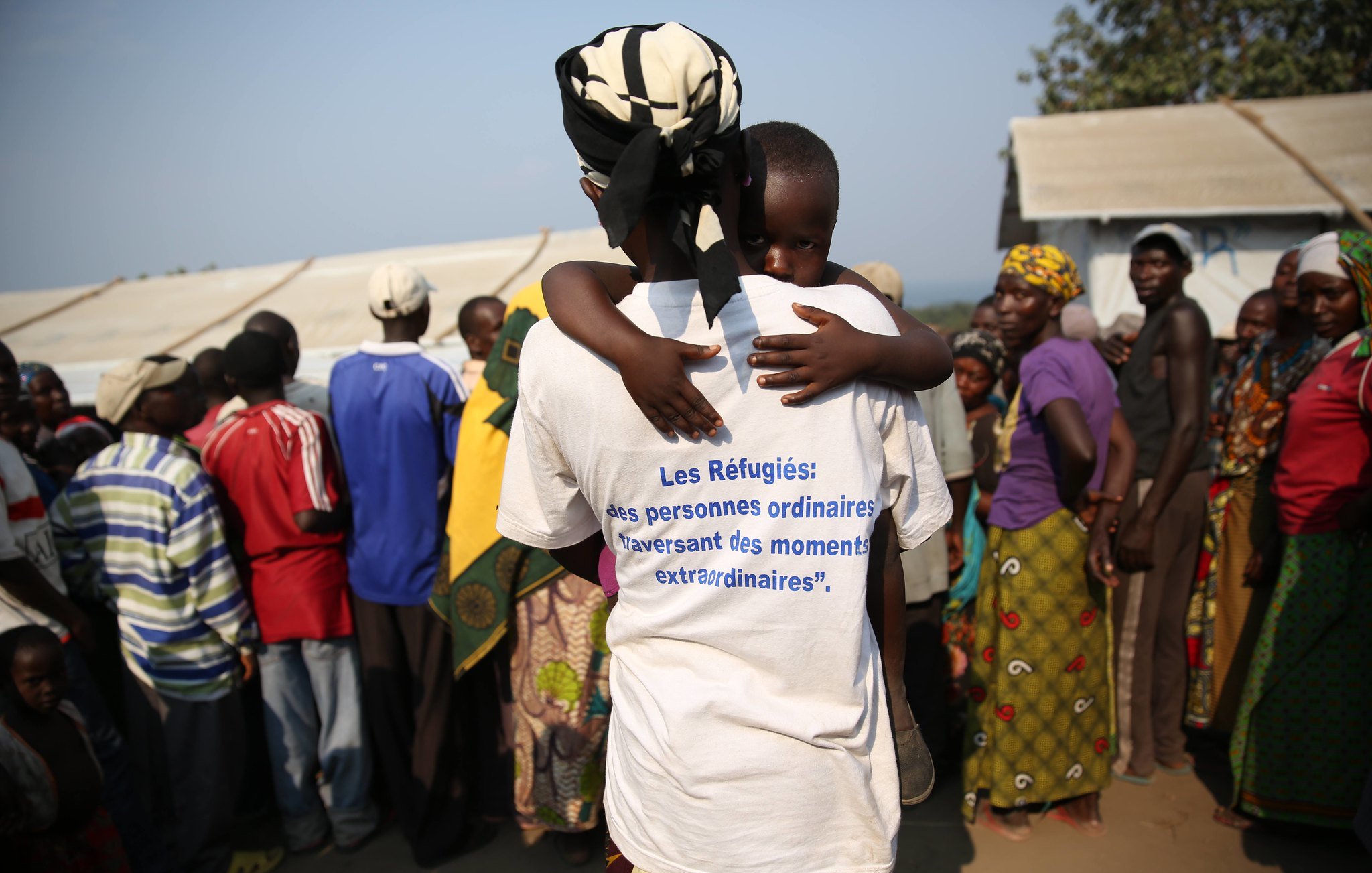 L'image montre un rassemblement de personnes dans un environnement de camp. Au premier plan, une femme, vue de dos, porte un t-shirt blanc portant une inscription en bleu qui dit : « Les Réfugiés : des personnes ordinaires traversant des moments extraordinaires. » Dans ses bras, elle tient un enfant, qui semble se blottir contre elle. En arrière-plan, on peut apercevoir un groupe de personnes, principalement des adultes, ayant des expressions variées. L'ensemble de la scène évoque des thèmes de vulnérabilité et de solidarité. Les vêtements des personnes présentes sont colorés, et l'atmosphère semble à la fois tendue et solidaire. Le décor suggère un campement temporaire, probablement en réponse à une crise.
