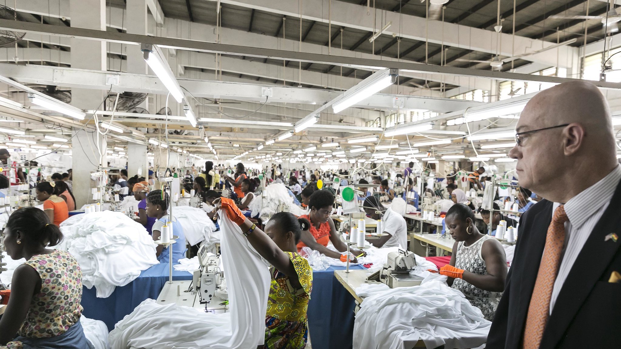 L'image montre une grande usine de couture animée. De nombreux ouvriers, principalement des femmes, sont occupés à coudre des vêtements, en particulier des chemises blanches. Ils travaillent en équipe autour de tables de couture, utilisant des machines à coudre. L'ambiance est dynamique, avec des mouvements rapides des mains et des tissus qui flottent. À droite, un homme en costume observe la scène, se tenant debout et faisant face au travail en cours. L'usine est bien éclairée, avec des lumières suspendues au plafond, et les murs sont de couleur claire, créant un espace de travail ouvert et organisé.