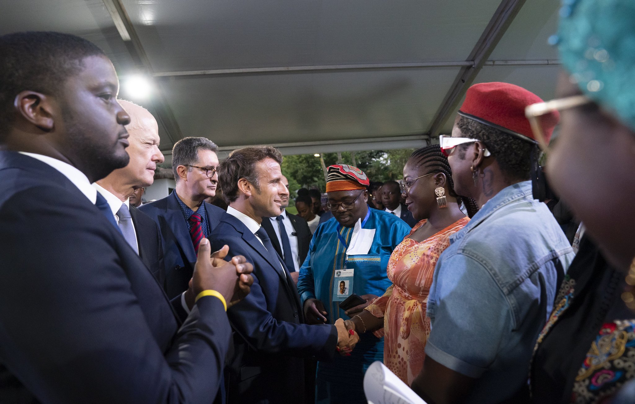 L'image représente un groupe de personnes réunies sous un chapiteau. Au centre, un homme en costume sombre, probablement un leader ou une personnalité importante, est en train de serrer la main d'une femme habillée de façon colorée. On peut apercevoir des visages souriants et une ambiance chaleureuse. D'autres personnes, habillées dans des tenues variées, entourent le couple, regardant avec intérêt. Les arrière-plans sont légèrement flous, mais on ressent une atmosphère de camaraderie et d'échange.