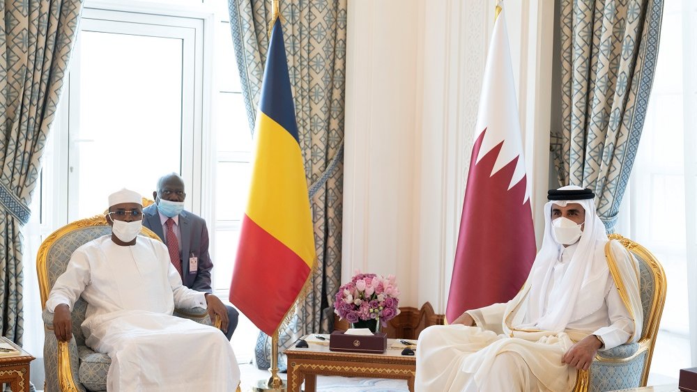 L'image montre une réunion formelle entre deux dirigeants. À gauche, un homme porte une tenue traditionnelle blanche et un chapeau, symbole de son pays. À sa droite, un autre homme est habillé d'une robe longue beige et d'un couvre-chef traditionnel. Entre eux, une table basse en bois contient des objets décoratifs. En arrière-plan, on peut voir des drapeaux : celui du Tchad à gauche et celui du Qatar à droite. La décoration des murs et des rideaux est élégante, avec des motifs riches et des couleurs apaisantes. L'atmosphère est solennelle et professionnelle.