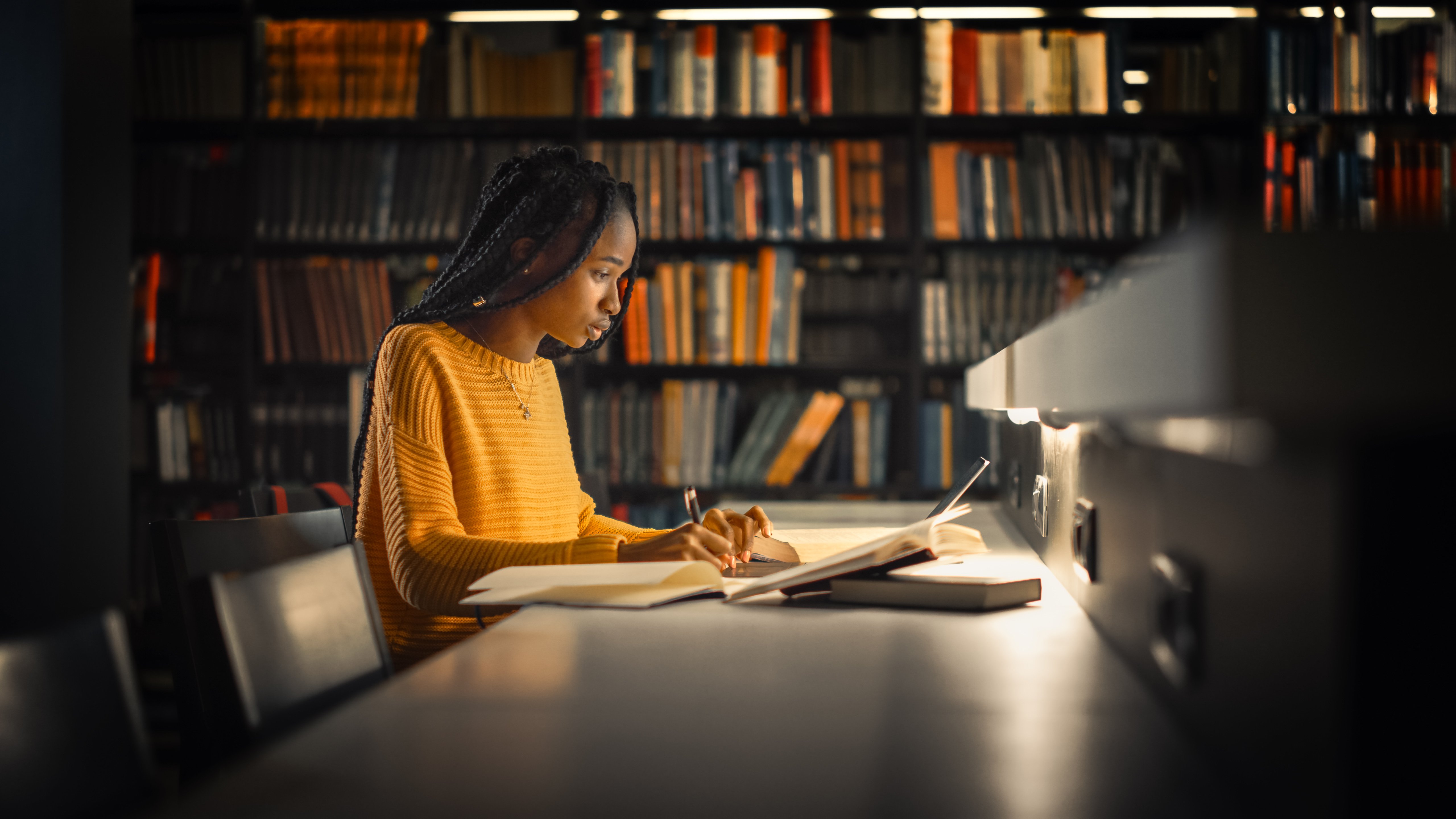 L'image montre une jeune femme assise à une table dans une bibliothèque. Elle est concentrée sur ses études, écrivant dans un cahier avec un stylo. Elle porte un pull en laine de couleur jaune. La lumière douce provenant d'une lampe de bureau éclaire son visage et son cahier. En arrière-plan, on voit des étagères remplies de livres, créant une atmosphère studieuse et calme, idéale pour la lecture et l'apprentissage. La posture de la jeune femme montre qu'elle est absorbée par son travail, avec une expression de détermination sur son visage.