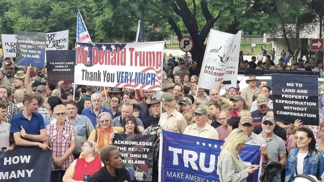 L'image montre une grande foule rassemblée lors d'une manifestation. Les participants tiennent divers panneaux et banderoles. Certains panneaux expriment leur soutien à Donald Trump avec des messages comme "Thank you VERY MUCH" et "MAKE AMERICA GREAT AGAIN". D'autres affichent des revendications ou des slogans liés à des préoccupations politiques. Les manifestants présentent une variété d'âges et de styles vestimentaires, certains portant des chapeaux emblématiques. L'atmosphère semble animée, avec des expressions de passion sur leurs visages. En arrière-plan, des arbres et des panneaux de signalisation sont visibles, créant un cadre urbain pour cette manifestation.