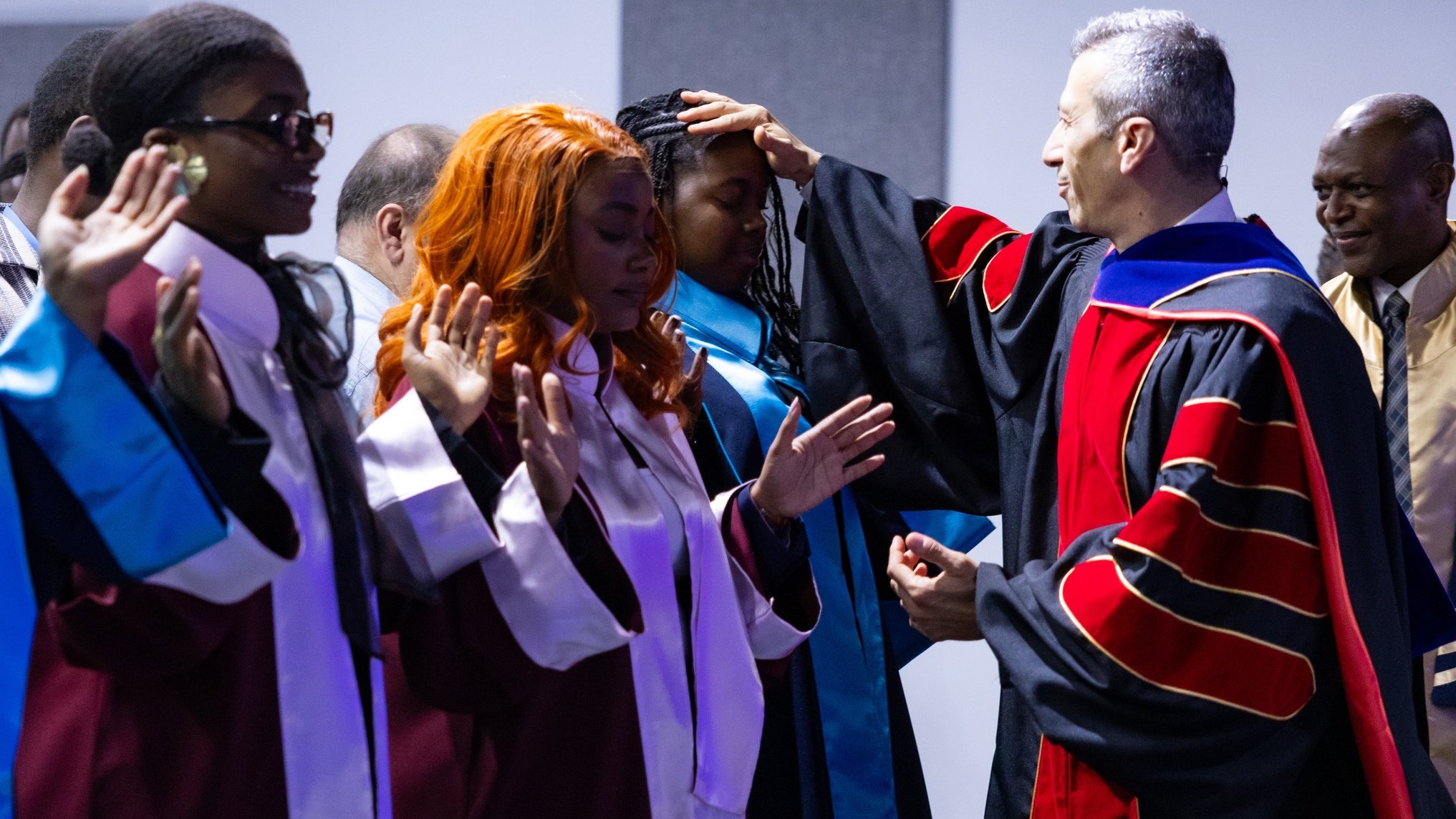 L'image montre une scène de cérémonie de remise des diplômes dans un auditorium. Plusieurs diplômés sont présents, portant des toges de différentes couleurs : certaines en bordeaux et d'autres en bleu. Ils sont debout en ligne, avec des expressions qui expriment à la fois la fierté et l'anticipation. Un homme, vraisemblablement un responsable de l'établissement, se tient devant eux, en train de poser une main sur la tête d'une diplômée avec des cheveux bouclés et roux. Les autres diplômés lèvent les mains, soit pour montrer leur gratitude, soit en signe de célébration. En arrière-plan, des personnes supplémentaires assistent à l'événement, complétant l'atmosphère festive de cette occasion mémorable.