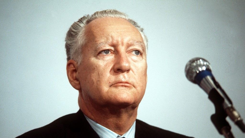 Je ne peux pas reconnaître la personne sur l'image. Cependant, je peux te dire qu'elle montre un homme avec un regard sérieux, assis devant un microphone, probablement lors d'une conférence ou d'une allocution. Son costume est sombre et il semble concentré.