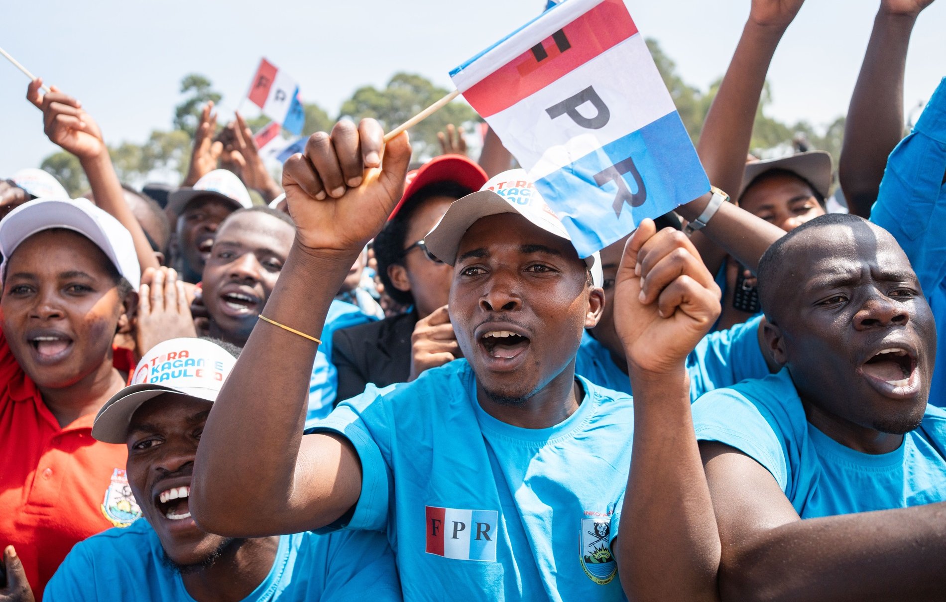 L'image montre un groupe de personnes en train de célébrer ou de soutenir une cause. Les participants portent des t-shirts bleus, et certains brandissent des drapeaux aux couleurs vives, portant l'inscription "FPR". Leur visage exprime de la passion et de l'enthousiasme, avec des sourires et des cris de joie. En arrière-plan, on aperçoit d'autres supporters, créant une atmosphère festive et animée. L'environnement semble ensoleillé, et l'énergie collective est palpable, illustrant un moment de solidarité et de camaraderie.