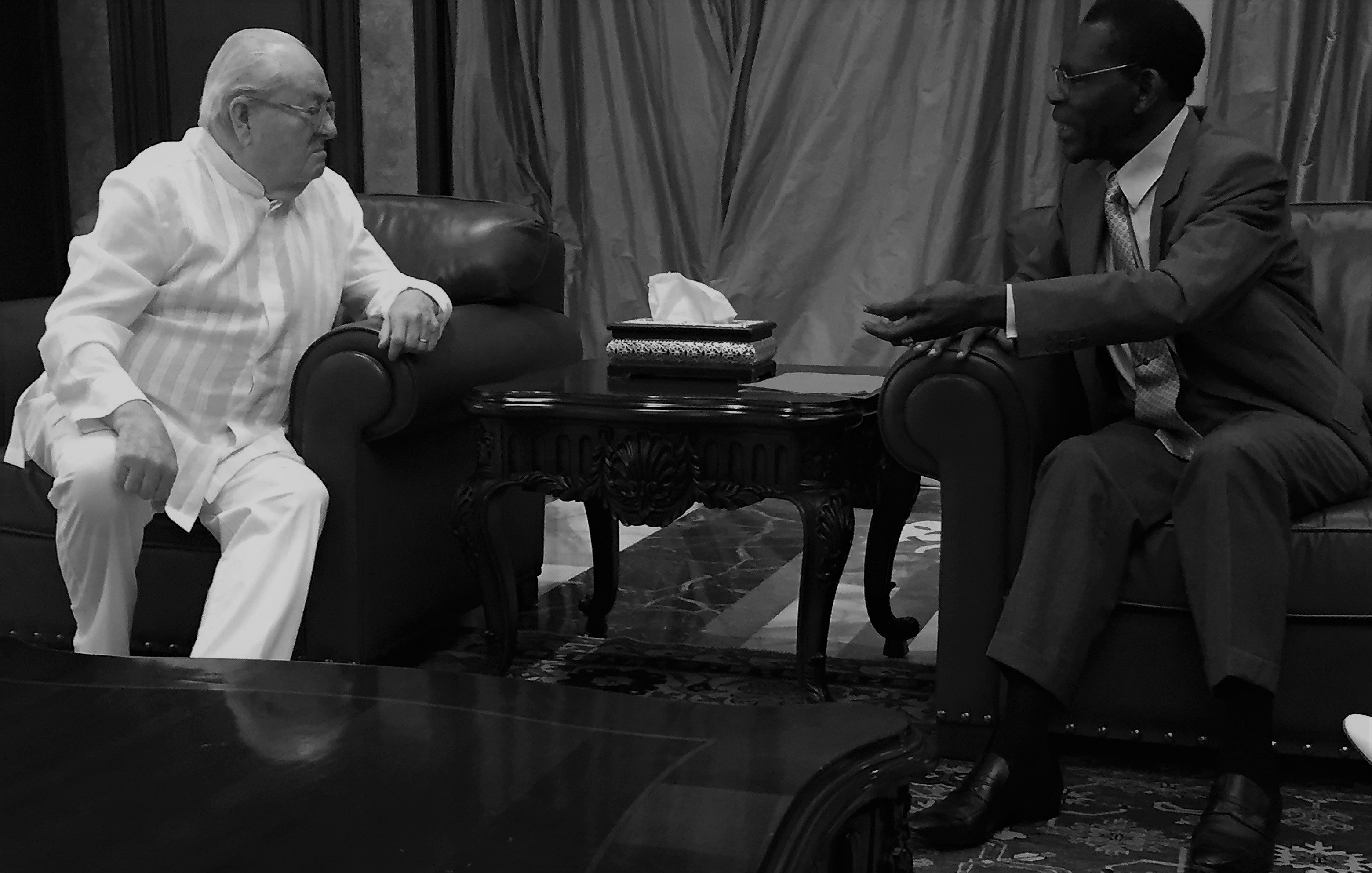 L'image représente une scène de discussion entre deux personnes assises l'une en face de l'autre dans un salon élégant. L'une des personnes porte un costume clair, tandis que l'autre est habillé d'un costume sombre. Ils sont assis sur des fauteuils en cuir, avec une table basse en bois ornée entre eux, où se trouve un objet décoratif. L'arrière-plan est composé de rideaux drapés et de murs en ton neutre, créant une atmosphère formelle et chaleureuse. L'éclairage est doux, ajoutant une sérénité à leur échange. On peut imaginer qu'ils discutent de manière engagée, leurs gestes accentuant l'intensité de la conversation.