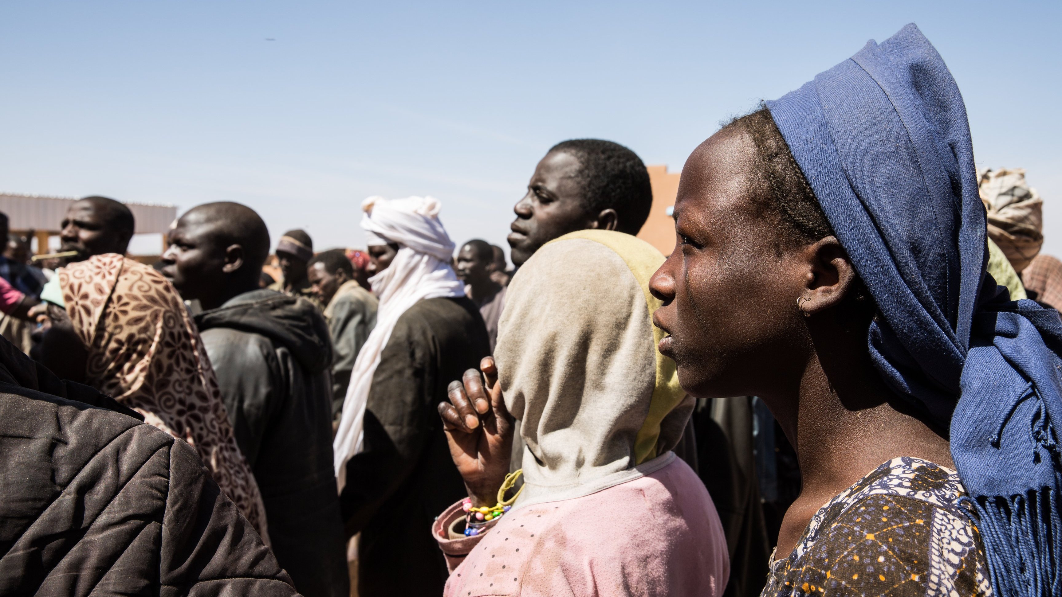 L'image montre un groupe de personnes se tenant debout, la plupart tournées vers un même point. On peut distinguer des hommes et des femmes de différents âges, habillés de vêtements variés, souvent colorés et traditionnels. Certaines personnes portent des couvertures sur la tête ou des scarves. L'expression sur leurs visages semble sérieuse, indiquant peut-être une attente ou une attention particulière. L'arrière-plan présente un ciel clair, suggérant une journée ensoleillée, et des structures émergent faiblement en arrière-plan, ajoutant à l'environnement d'un rassemblement. Le moment semble chargé d'émotions et de tension collective.