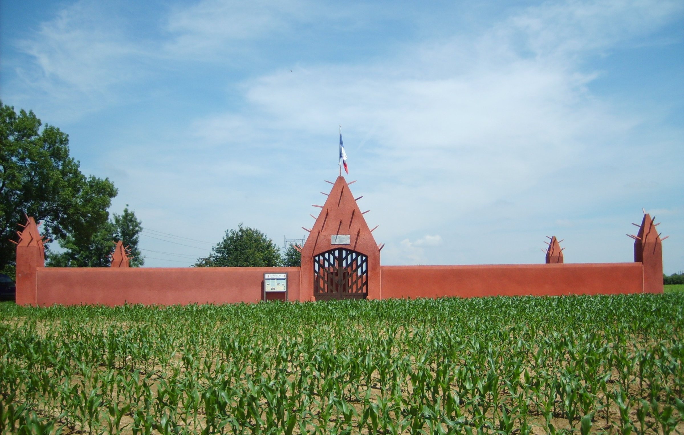 L'image présente une grande entrée de style architectural distinctif, peinte en rouge, qui s'élève au-dessus d'un champ de maïs. La structure est dotée d'un mur haut, orné de pointes qui se projettent vers le ciel, créant un aspect à la fois imposant et décoratif. Au centre, une porte en fer forgé permet d'entrer, et on peut apercevoir un drapeau français flottant au-dessus. Le ciel d'un bleu éclatant, parsemé de nuages légers, contraste avec la verdure des jeunes plants de maïs qui s'étendent devant le mur, ajoutant une touche de fraîcheur à la scène.
