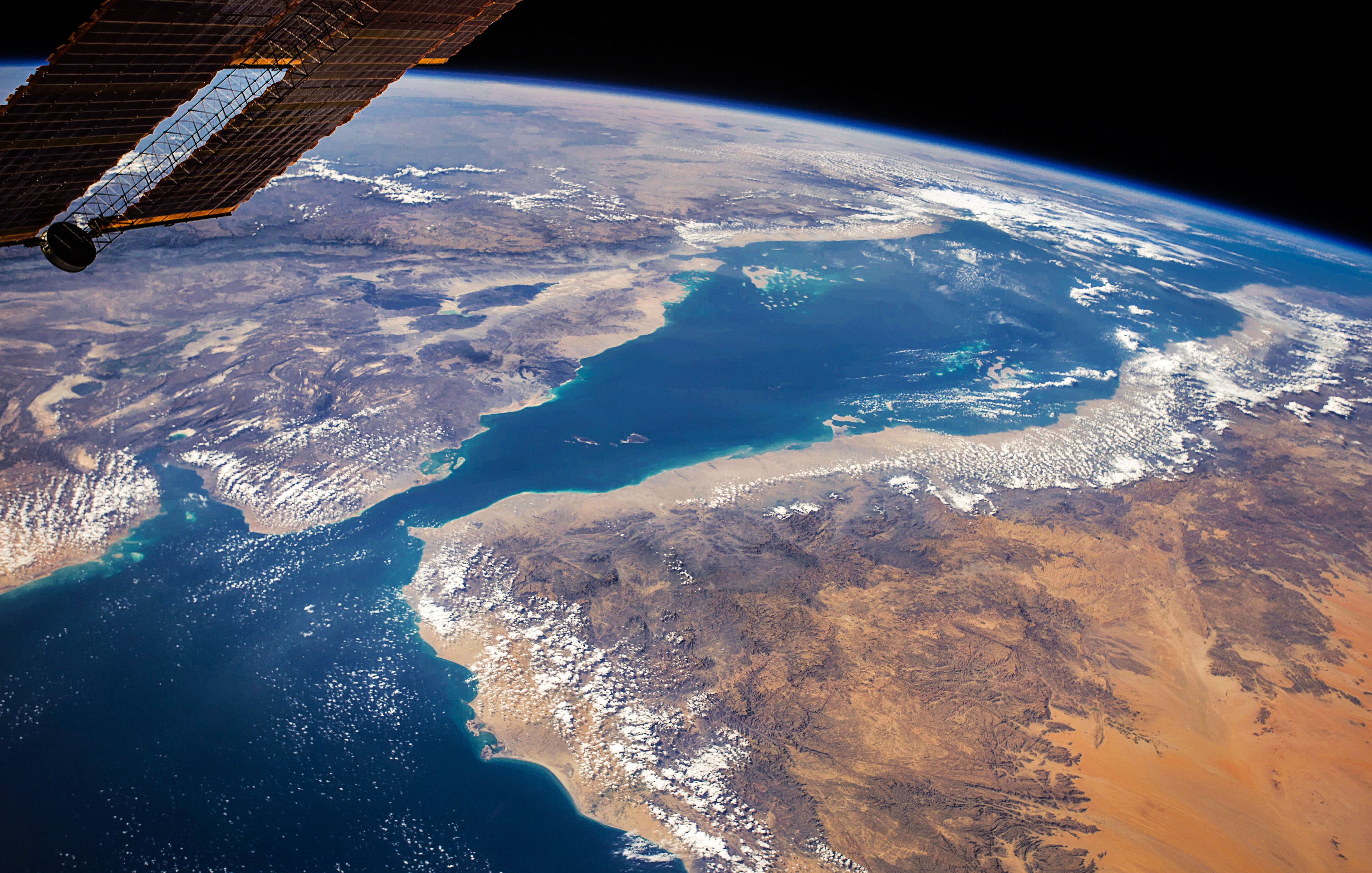 Cette image, prise depuis l'espace, montre une vue spectaculaire de la Terre. On y voit des paysages variés, avec une mer d'un bleu profond bordée de terres aux couleurs chaudes, tantôt sableuses, tantôt montagneuses. Il y a des nuages éparpillés qui ajoutent du relief à la scène. Les contours des côtes et des îles se dessinent nettement. En haut de l'image, on peut apercevoir la noirceur de l'espace, ce qui crée un beau contraste avec les teintes vives de la planète. Cette vue donne une impression de sérénité et d'immensité, rappelant la beauté fragile de notre environnement.