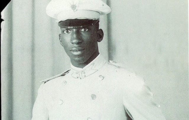 L'image montre un jeune homme en tenue militaire. Il porte un uniforme blanc qui se compose d'une chemise et d'un pantalon assortis, avec une ceinture qui souligne sa taille. Sur sa tête, il a une casquette blanche ornée d'un insigne. Son expression est sérieuse et déterminée. L'arrière-plan est neutre, probablement un drap ou un fond uni, ce qui met en valeur sa silhouette et son uniforme.