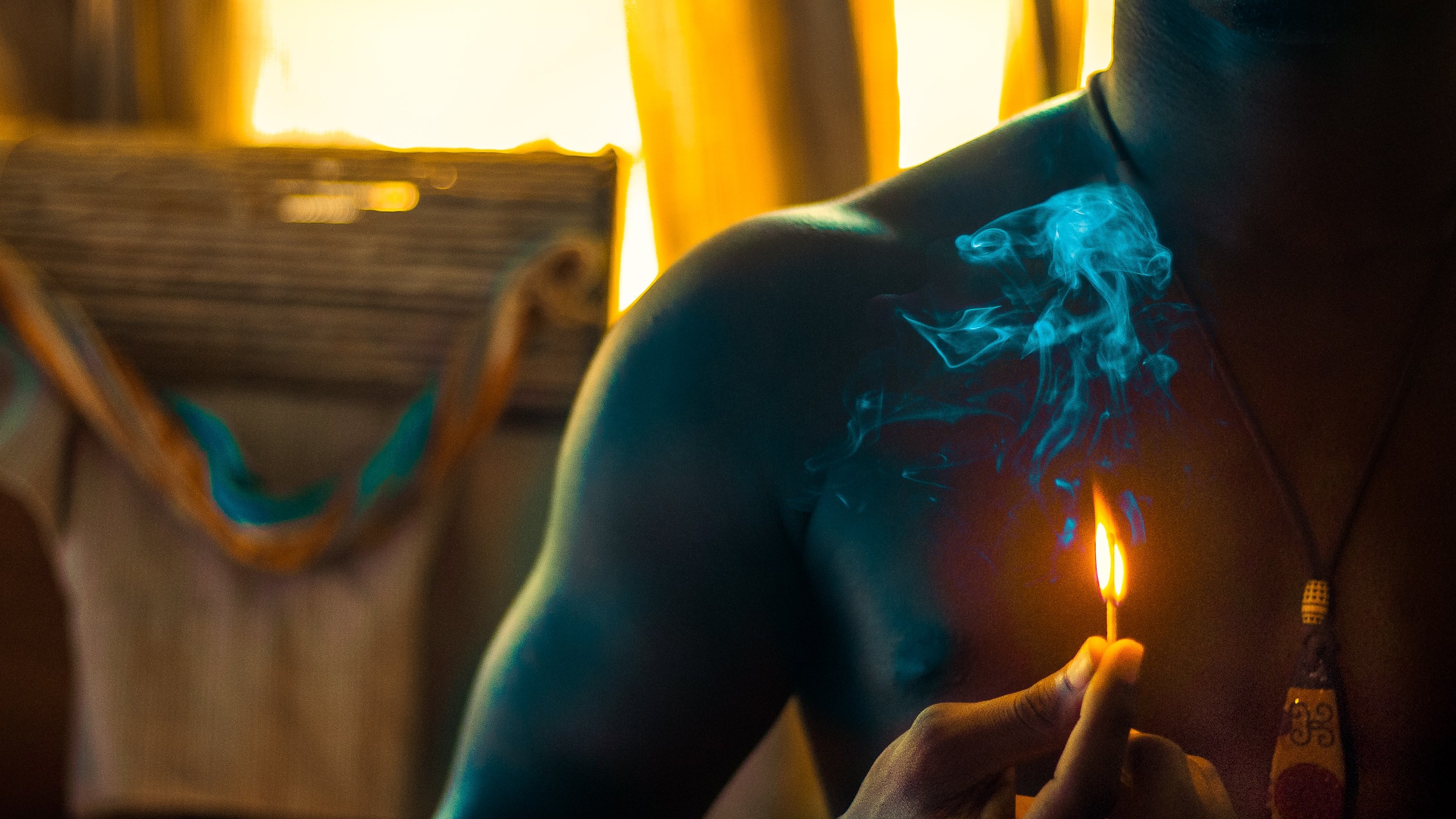 Dans cette image, nous voyons un homme torse nu tenant une petite flamme d'une allumette qui fume légèrement. La lumière douce qui l'entoure crée une ambiance chaleureuse. À l'arrière-plan, des éléments flous suggèrent un environnement intérieur, peut-être une pièce éclairée par le soleil couchant, ce qui ajoute une touche de chaleur à l'atmosphère. La fumée s'élève lentement, créant un contrasté avec la lueur de la flamme.