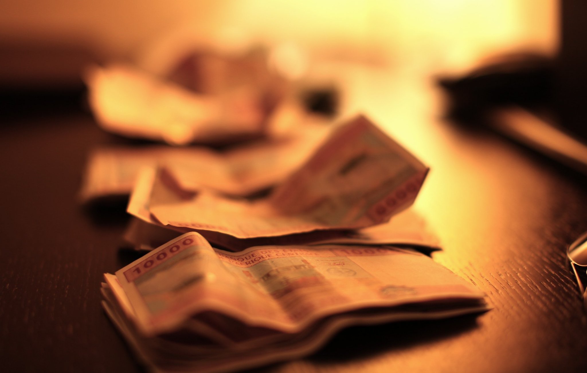 L'image montre un plan rapproché de billets de banque étalés sur une surface sombre, probablement une table en bois. Les billets sont flous, suggérant une légère profondeur de champ, ce qui attire l'attention sur la texture et les détails des billets. L'éclairage est doux et chaud, avec une lumière diffuse provenant d'une source hors du champ de vision, créant une ambiance intime et feutrée. Cette lumière fait ressortir les motifs et les couleurs des billets, ajoutant une dimension visuelle à l'ensemble.