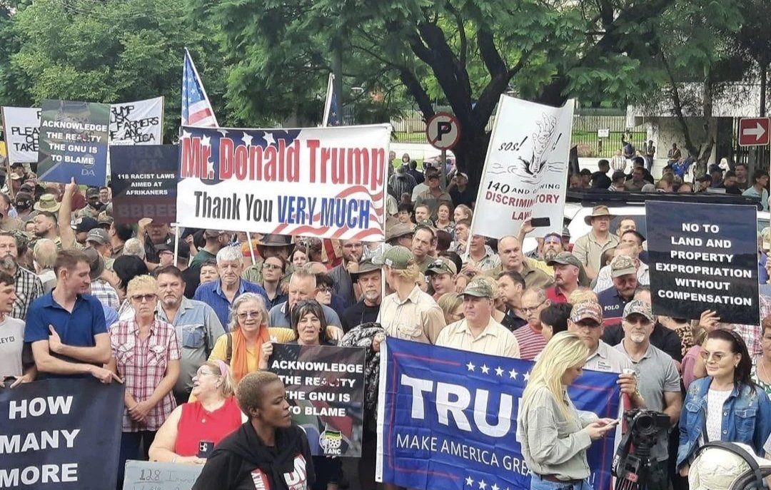 L'image montre une grande foule rassemblée lors d'une manifestation. Les participants tiennent divers panneaux et banderoles. Certains panneaux expriment leur soutien à Donald Trump avec des messages comme "Thank you VERY MUCH" et "MAKE AMERICA GREAT AGAIN". D'autres affichent des revendications ou des slogans liés à des préoccupations politiques. Les manifestants présentent une variété d'âges et de styles vestimentaires, certains portant des chapeaux emblématiques. L'atmosphère semble animée, avec des expressions de passion sur leurs visages. En arrière-plan, des arbres et des panneaux de signalisation sont visibles, créant un cadre urbain pour cette manifestation.