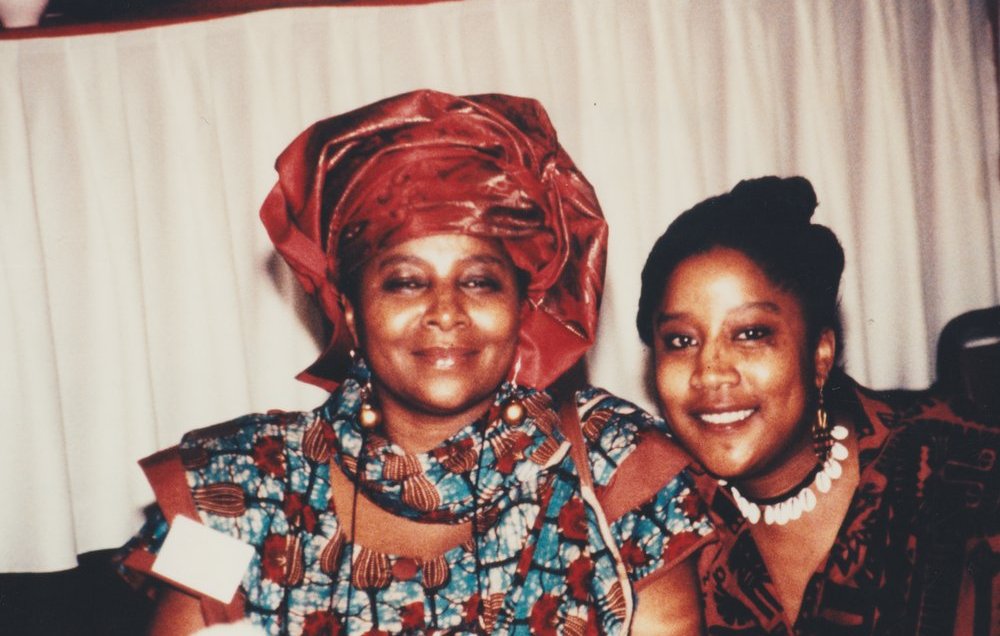 L'image montre deux femmes souriantes posant côte à côte. La femme à gauche porte un vêtement traditionnel aux motifs colorés, accompagné d'un foulard assorti. Sa expression est douce et chaleureuse. À sa droite, la seconde femme affiche également un grand sourire ; elle porte un haut à motifs et des accessoires qui ajoutent une touche élégante à son apparence. L'environnement semble être festif, avec une nappe blanche en arrière-plan, laissant planer une atmosphère accueillante et joyeuse.