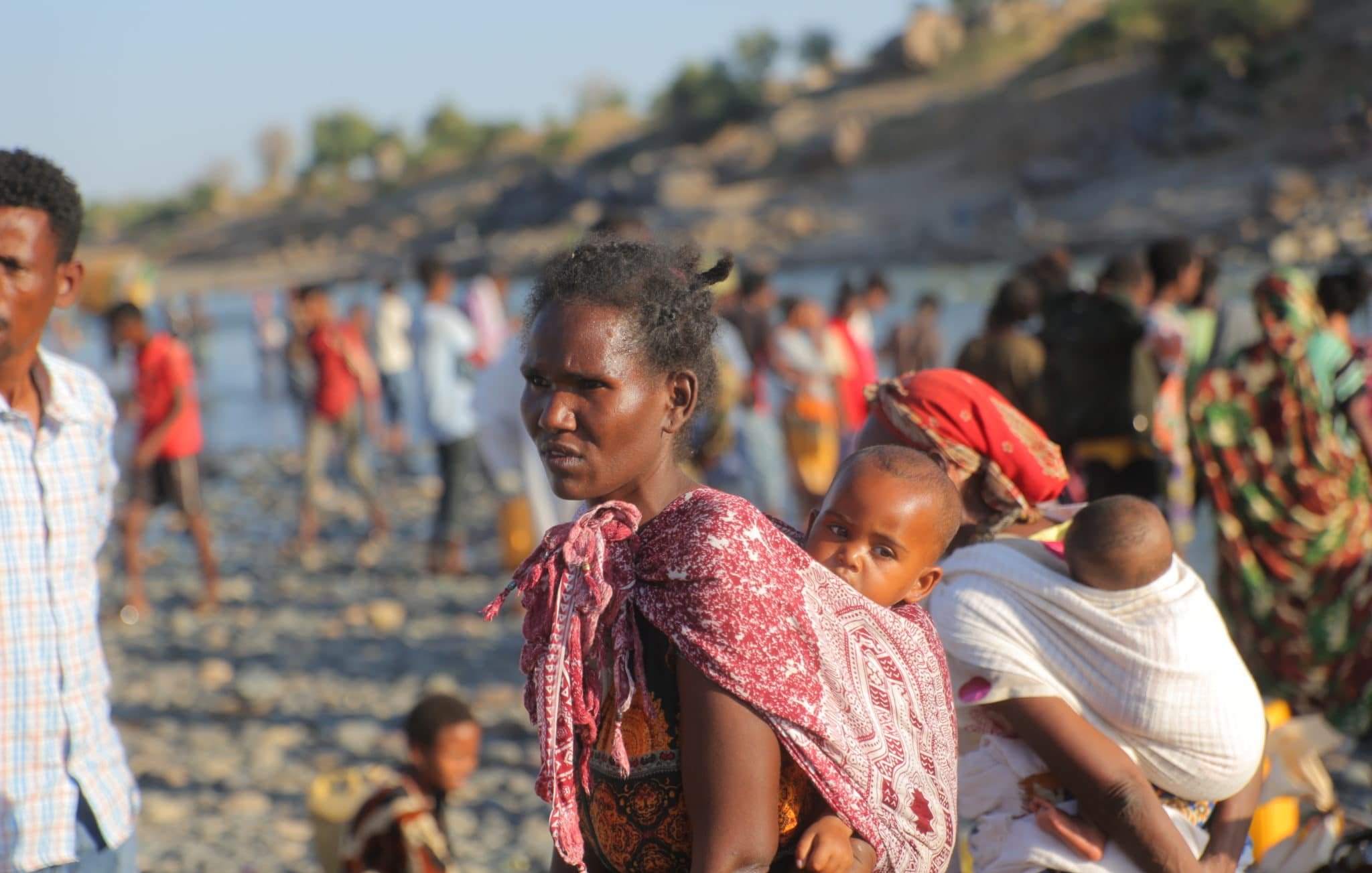 L'image représente une scène vibrante et émotive d'un groupe de personnes au bord d'une rivière. Au premier plan, une femme porte un enfant sur son dos, enveloppé dans un tissu coloré. Son expression est sérieuse et déterminée. Elle est entourée d'autres personnes, dont certaines portent également des enfants. Les vêtements des personnes sont variés, avec des motifs et des couleurs vives. En arrière-plan, on aperçoit des rochers et une végétation légère, ainsi que d'autres silhouettes de personnes se déplaçant le long de la rive. L'atmosphère semble chargée d'une mixité d'activités et d'émotions, reflétant à la fois la vie quotidienne et des moments de transition.