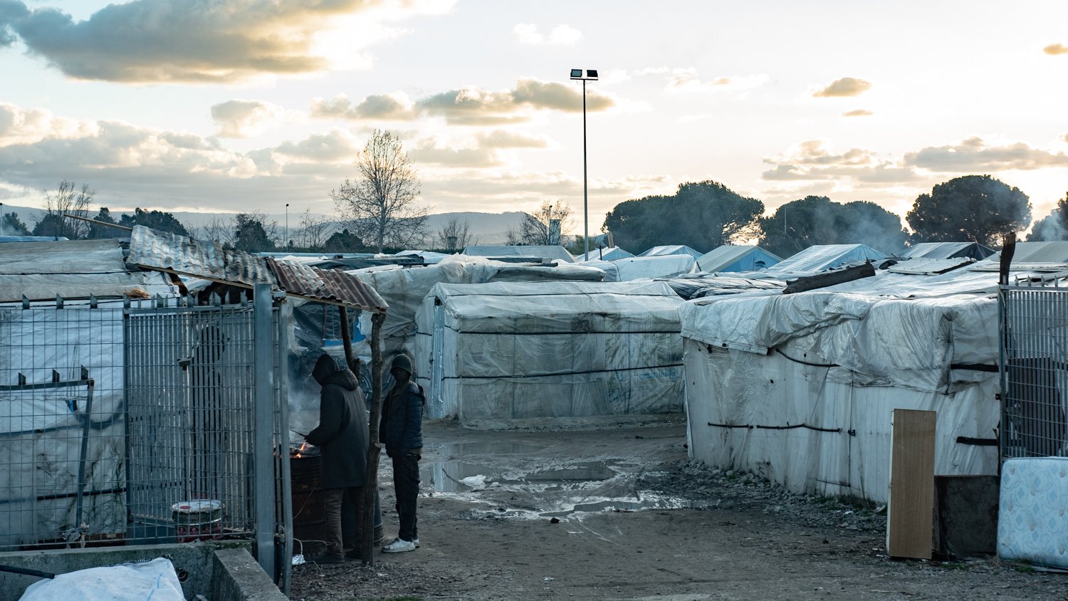 L'image montre un paysage de campement, probablement un bidonville, où des abris temporaires sont installés. Ces structures, couvertes de bâches blanches, sont disposées de manière désordonnée, formant des allées étroites entre elles. Au premier plan, deux personnes se tiennent près d'un feu, suggérant un moment de convivialité ou de chaleur dans un environnement difficile. Le ciel est nuageux, avec des teintes de bleu et d'orange, indiquant une heure de la journée où le soleil se lève ou se couche. En arrière-plan, des arbres et des monts se dessinent, ajoutant une dimension naturelle à ce paysage urbain. L'atmosphère peut sembler mélancolique, mais aussi résiliente, avec des traces de vie et d'activités humaines.