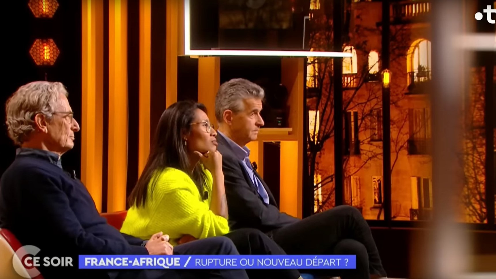 L'image montre un plateau de télévision lors d'une émission intitulée "France-Afrique : Rupture ou nouveau départ ?". Trois personnes sont assises sur un canapé, éclairées par des lumières chaudes. À gauche, un homme avec des lunettes et des cheveux grisonnants porte un pull sombre. Au centre, une femme, habillée d'un pull lumineux, semble attentive et réfléchie. À droite, un homme aux cheveux courts et argentés affiche une expression sérieuse. En arrière-plan, on peut apercevoir des fenêtres illuminées qui donnent une ambiance chaleureuse à l'ensemble. Le décor est moderne, avec des éléments graphiques en arrière-plan qui ajoutent à l'esthétique de l'émission.