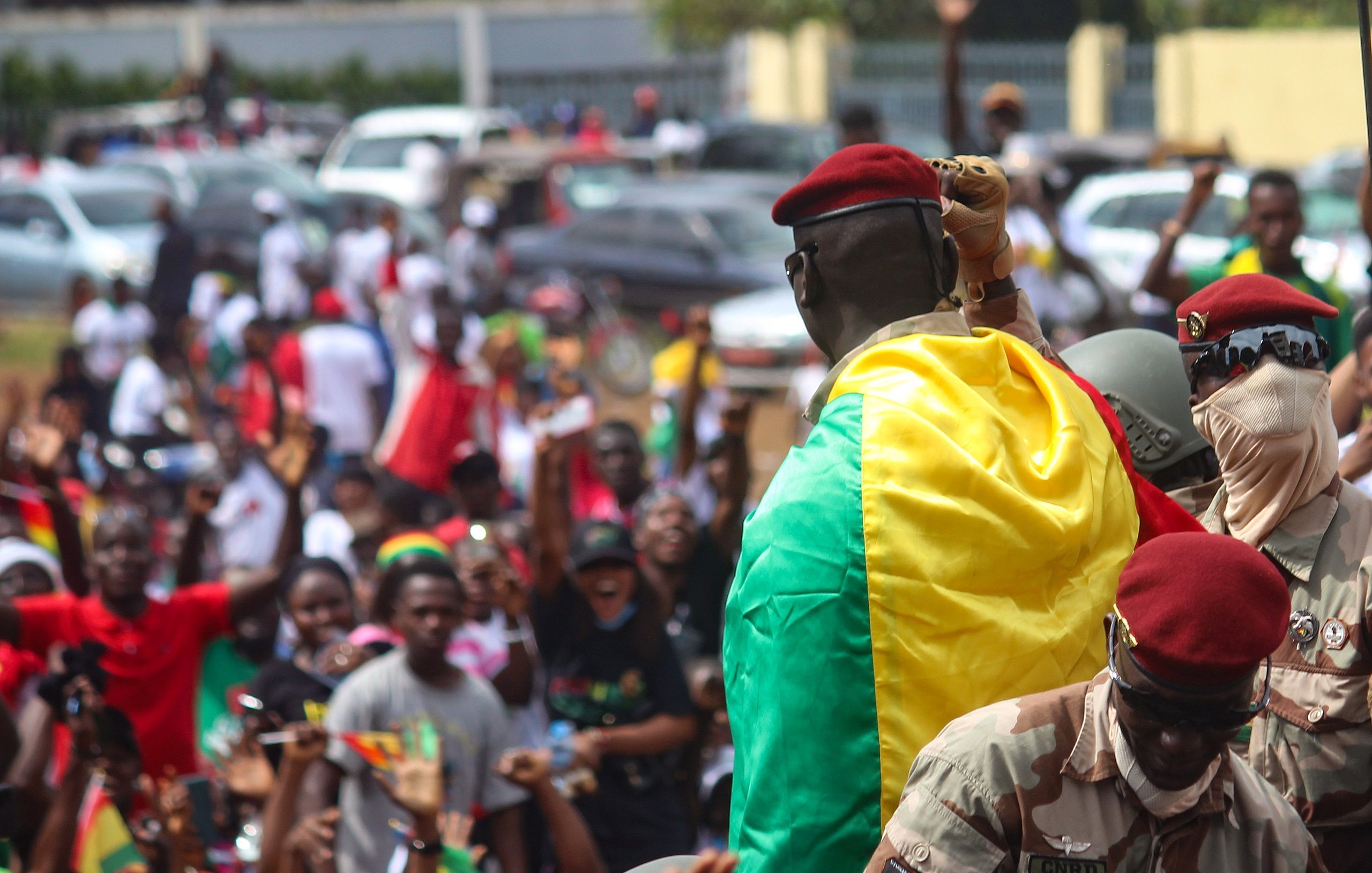 L'image montre une scène dynamique et pleine d'énergie. Au premier plan, un homme en uniforme militaire, portant un béret rouge, se tient sur une plateforme surélevée. Il est enveloppé dans un drapeau aux couleurs vives du Mali, qui est vert, jaune et rouge. Autour de lui, une foule enthousiaste s'agite, brandissant des drapeaux et exprimant leur joie. Les visages des personnes dans la foule montrent des émotions variées allant de l'excitation à la fierté. En arrière-plan, on peut voir des voitures stationnées et d'autres participants se mêlant dans une atmosphère de célébration collective. L'ensemble dégage un fort sentiment d'unité et de patriotisme.