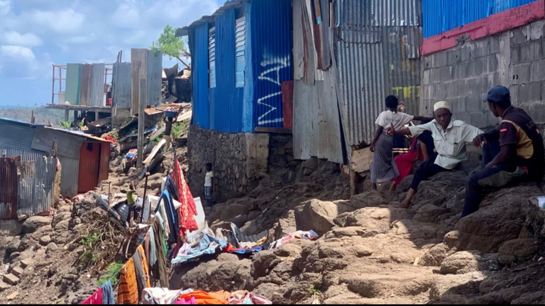Cette image présente un paysage d'un quartier informel, caractérisé par des habitations en tôle et en béton, construite sur un terrain escarpé. Les maisons, souvent de différentes couleurs, semblent inachevées avec des parois en métal ondulé. Au premier plan, des personnes sont assises sur des rochers, discutant ou s'affairant. Le sol est accidenté, recouvert de débris et de vêtements épars, tandis qu'en arrière-plan, des structures réduites à des vestiges montrent l'impact de l'environnement sur ce lieu de vie. Le ciel est clair, laissant présager une journée ensoleillée. L'atmosphère générale évoque une communauté résiliente, malgré les difficultés et les conditions de vie précaires.