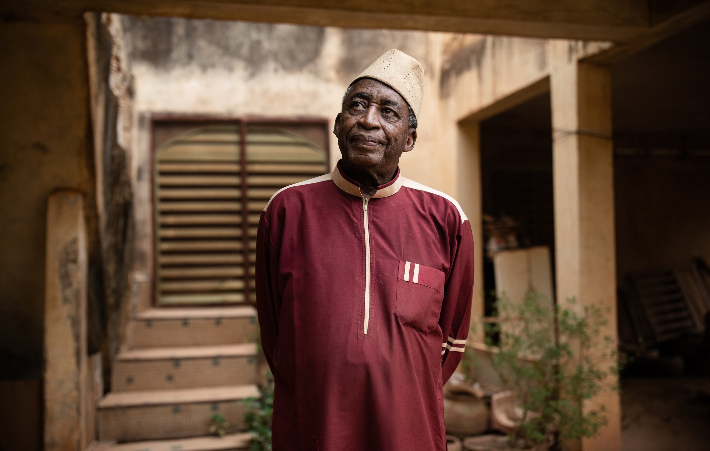 L'image montre un homme âgé portant une tenue traditionnelle d'une couleur rouge foncé. Sa chemise longue est agrémentée de détails blancs. Il se tient debout, les bras le long du corps, et regarde légèrement vers le haut. L'arrière-plan présente un bâtiment avec des murs usés et une porte en bois, ainsi que quelques plantes verdoyantes qui ajoutent une touche de nature à la scène. L'homme dégage une expression calme et réfléchie, créant une atmosphère de sagesse et de sérénité.