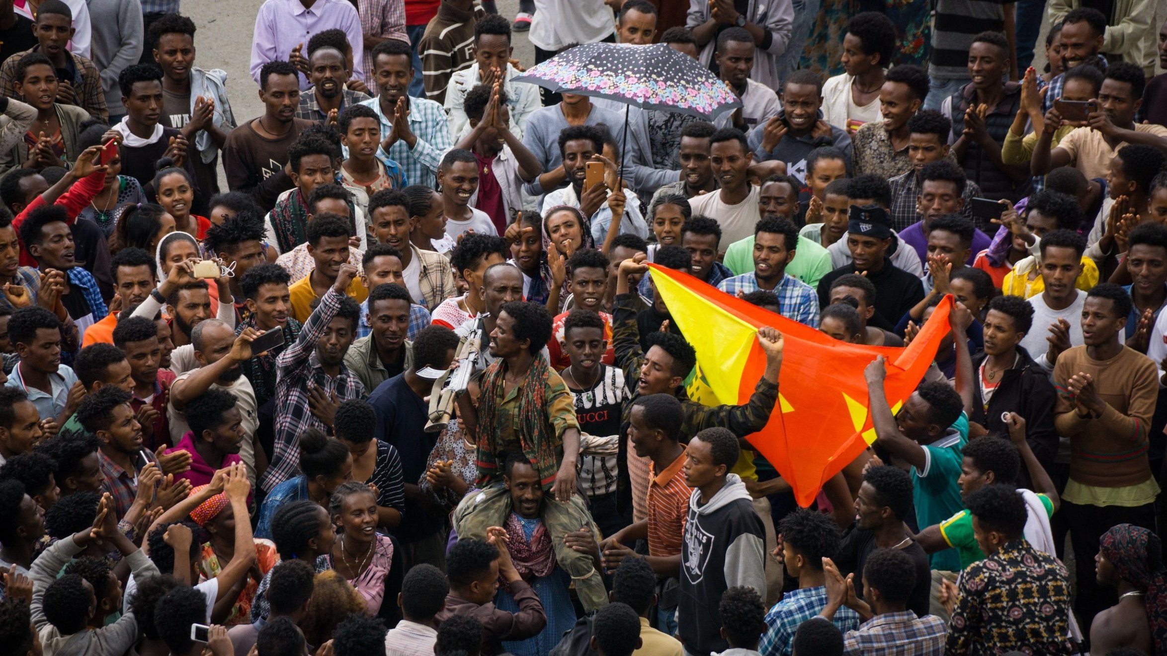 L'image représente une scène vibrante et animée d'une foule rassemblée ensemble. Au premier plan, on peut voir un groupe dense de personnes, principalement des jeunes hommes, exprimant leur énergie et leur enthousiasme. Certains d'entre eux lèvent les bras, d'autres applaudissent ou tiennent des drapeaux aux couleurs vives, notamment le rouge et le jaune. Au centre, une personne semble parler ou chanter, attirant l'attention de la foule. Les visages expriment des émotions variées telles que la joie, la passion et la détermination. Par temps peut-être nuageux, un parapluie coloré est en arrière-plan, ajoutant une note de couleur à la scène. L'ambiance générale est celle d'un rassemblement convivial, où les participants partagent un moment fort ensemble.