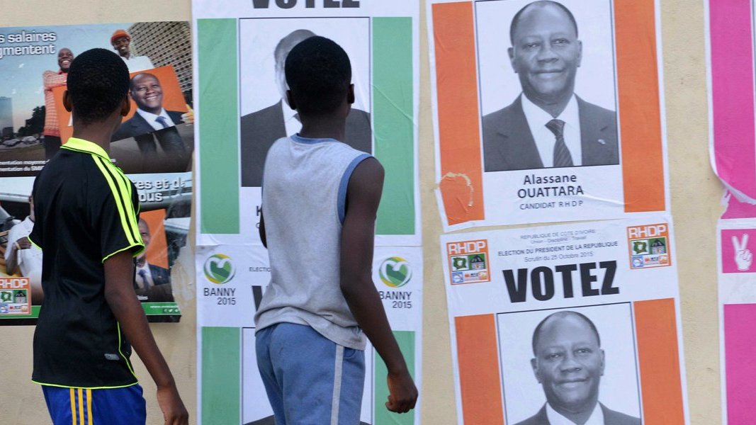 L'image montre un groupe de jeunes garçons marchant le long d'un mur recouvert d'affiches électorales. Les affiches présentent des portraits de candidats politiques, dont certaines affichent le nom d'Alassane Ouattara. Les affiches sont aux couleurs vives, avec des fonds colorés, principalement du rose et du vert, et des mots comme "VOTEZ". Les garçons, vêtus de t-shirts, semblent attentifs aux affiches. Le décor est urbain et évoque l'ambiance d'une campagne électorale animée.