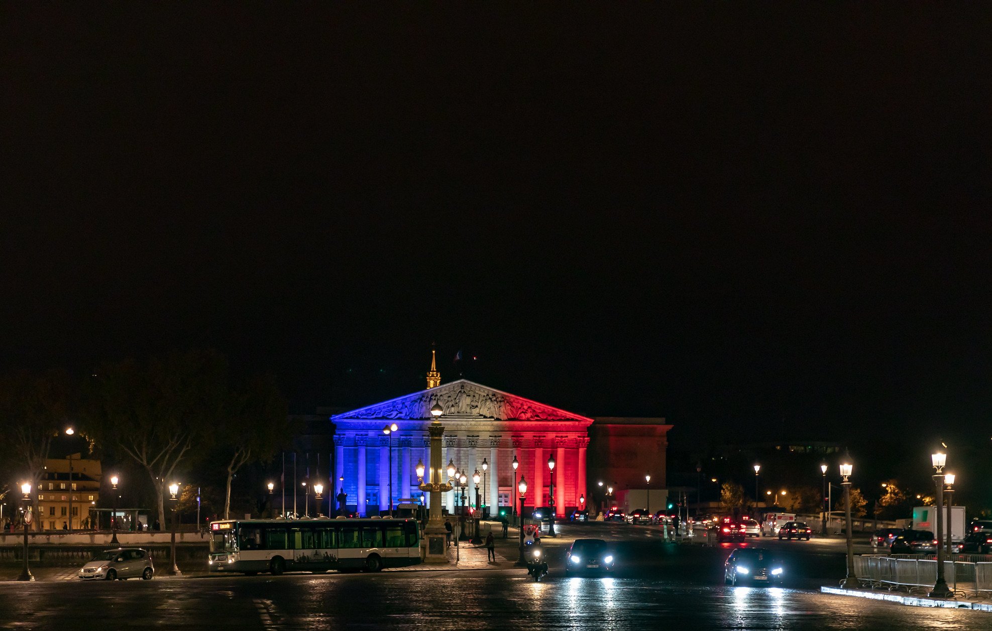 Imaginez un bâtiment majestueux illuminé par des lumières tricolores. La façade est peinte de bleu, de blanc et de rouge, évoquant les couleurs du drapeau français. Le ciel nocturne est sombre, créant un contraste saisissant avec les lumières vibrantes. À l'avant, vous pouvez sentir la présence d'une place animée, avec des voitures qui circulent et des lampadaires qui diffusent une lumière douce sur le pavé. La scène dégage une ambiance festive et patriotique, symbolisant la fierté nationale.