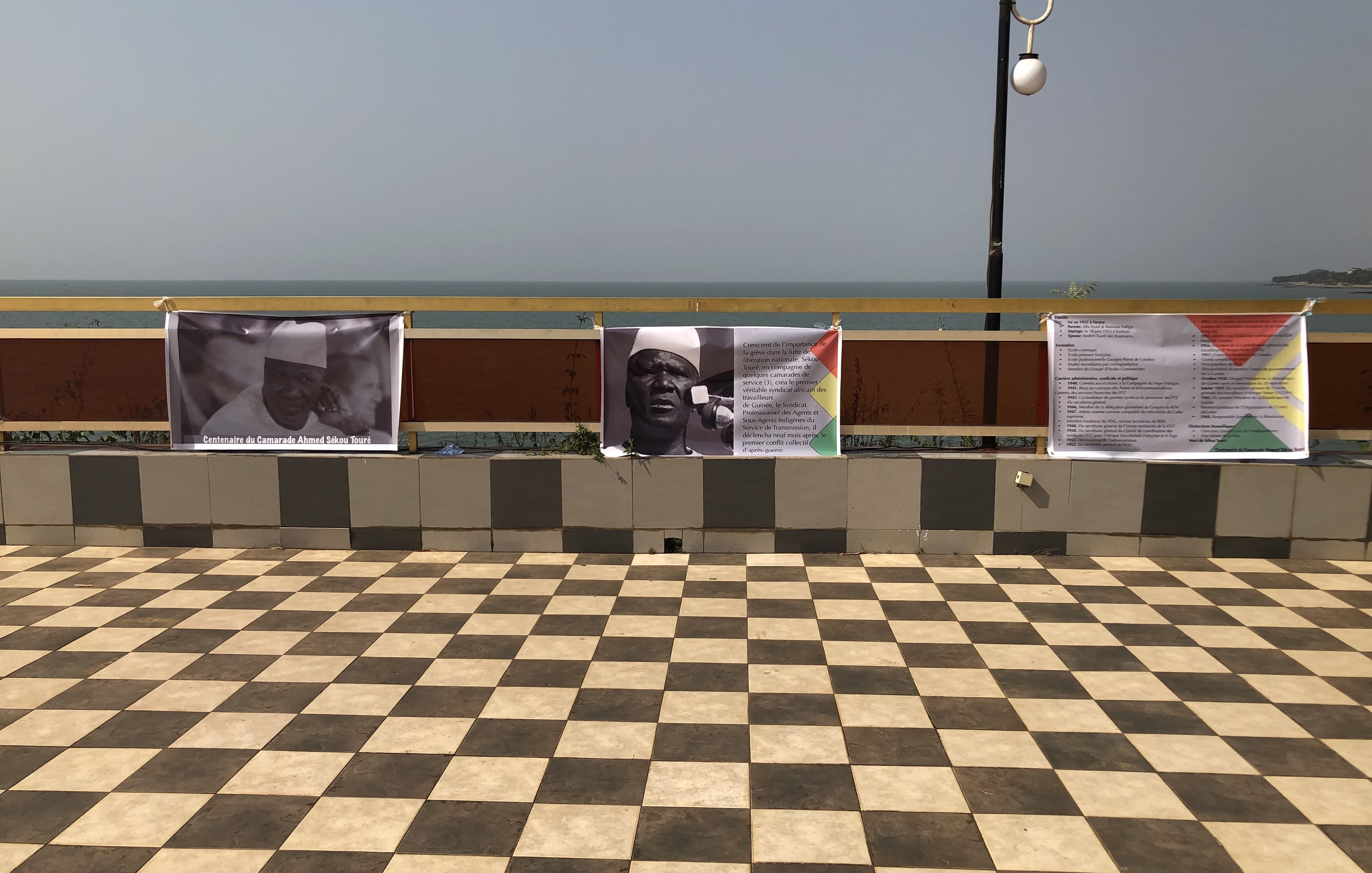 L'image montre un espace extérieur avec un sol en carreaux de damier, alternant entre des teintes claires et foncées. En arrière-plan, on aperçoit une étendue d'eau qui pourrait être la mer ou un lac, sous un ciel clair. Sur un muret, plusieurs affiches sont accrochées. Ces affiches semblent contenir du texte et des images en rapport avec un événement ou une exposition. L'environnement dégage une atmosphère calme, avec un éclairage naturel et une vue agréable sur l'eau.