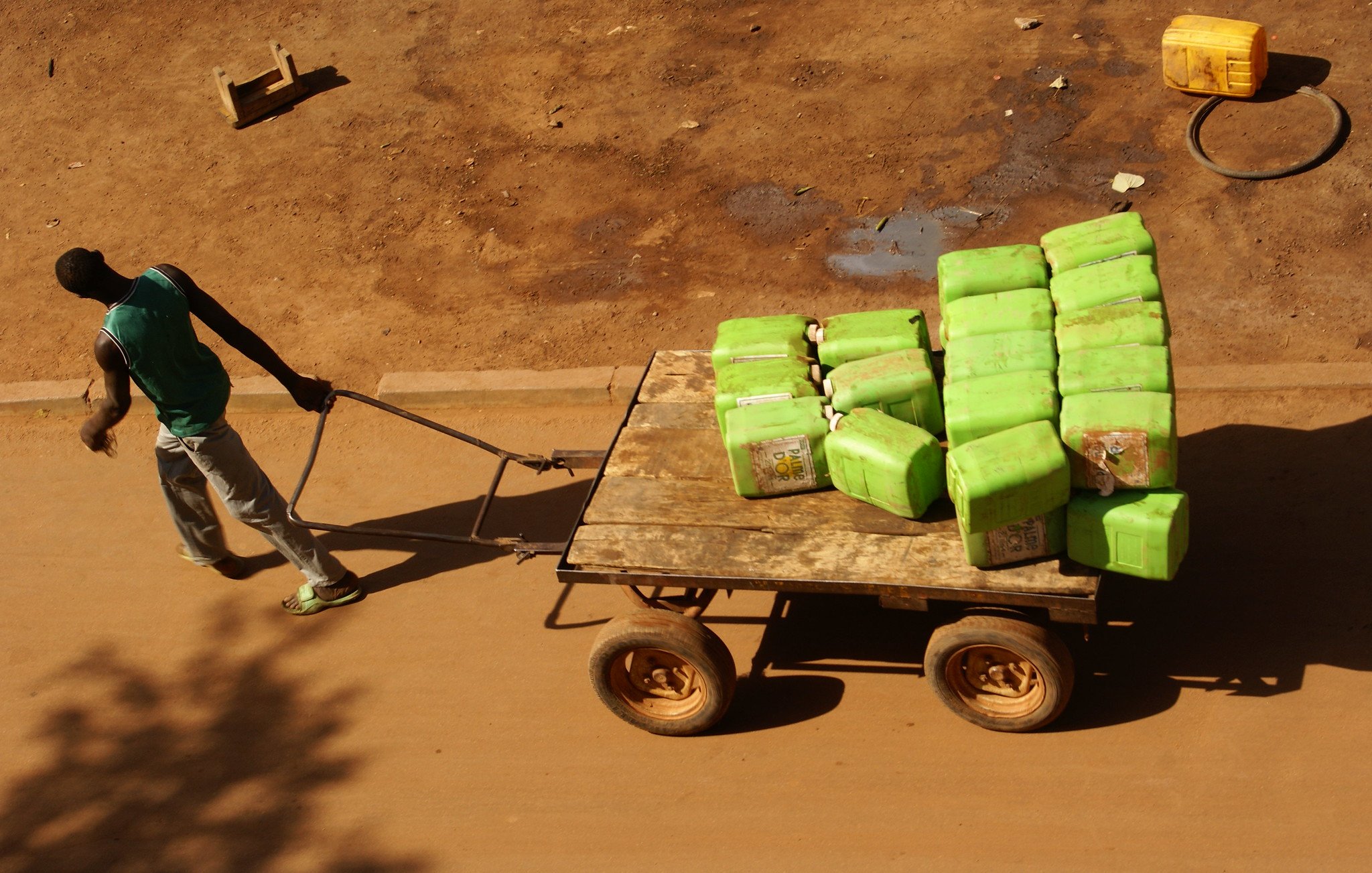 L'image montre un homme poussant une charrette en bois sur une route en terre. La charrette est remplie de nombreuses bouteilles vertes, probablement des contenants d'eau. L'homme porte un T-shirt sans manches et un pantalon gris, et son corps semble penché en avant, indiquant l'effort physique qu'il fournit. Le sol autour de lui est sec et légèrement poussiéreux, avec quelques traces visibles montrant que d'autres véhicules ou personnes ont déjà passé. En arrière-plan, il pourrait y avoir des objets comme des seaux ou des tuyaux, mais l'accent est mis sur l'homme et sa cargaison. La lumière est forte, suggérant une journée ensoleillée.