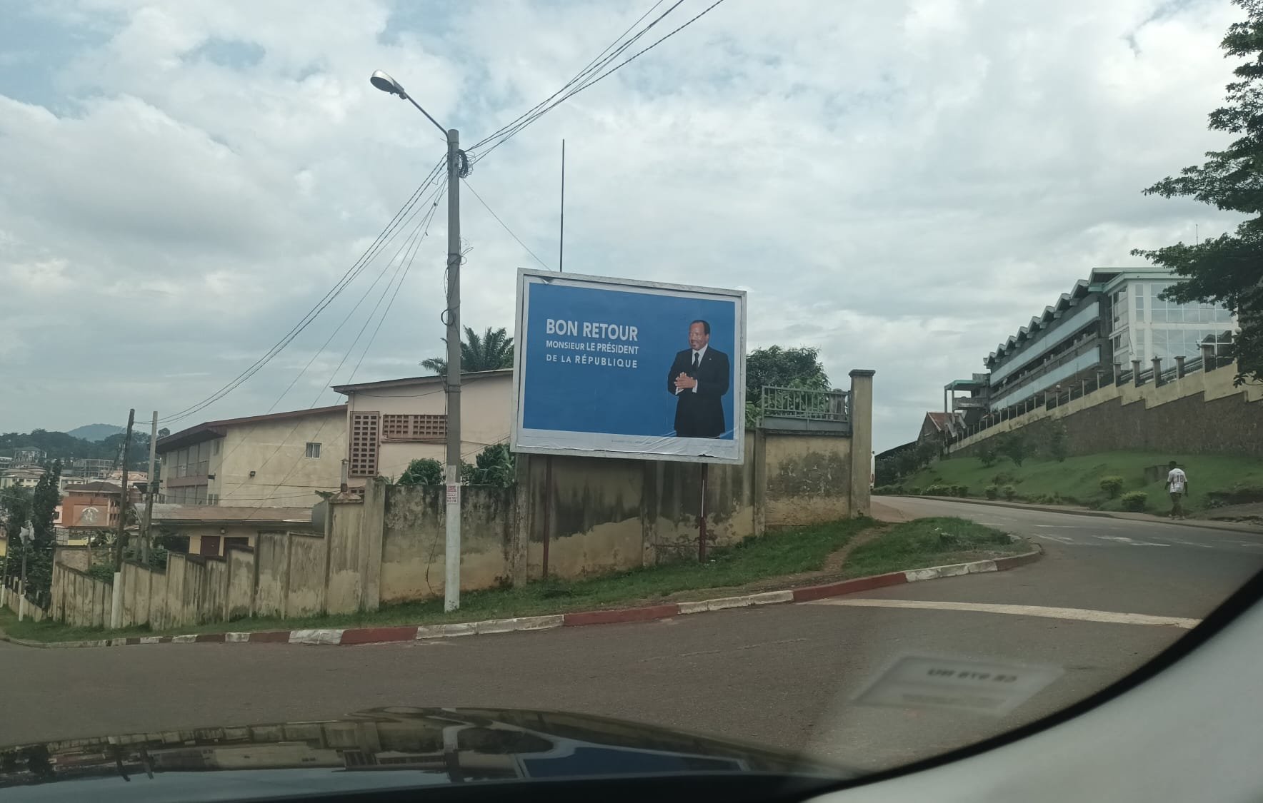 L'image montre une grande affiche publicitaire installée sur un mur à l'extérieur. Sur cette affiche, un homme, probablement un homme politique, se tient debout avec un sourire, vêtu d'un costume. Le fond est bleu, ce qui attire l'attention. En haut de l'affiche, on peut lire le texte "BON RETOUR", suivi d'un message qui semble faire référence à un événement ou une situation spécifique. En dessous, s'étend un paysage urbain avec des bâtiments, des routes et des arbres, et l'image suggère un périphérique ou une route en pente. Le ciel est partiellement nuageux, ajoutant une ambiance calme à la scène.