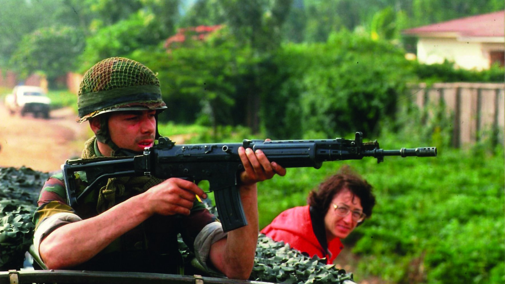 L'image montre un soldat, assis dans un véhicule militaire,tenant un fusil vers l'avant. Il est vêtu d'un uniforme camouflage et porte un casque en treillis. Son visage exprime la concentration et la vigilance, tandis qu'il regarde attentivement son environnement. En arrière-plan, une femme à lunettes observe la scène, semblant préoccupée. La verdure luxuriante entoure le cadre, créant un contraste avec la terre battue sur laquelle se trouve le véhicule. L'atmosphère de l'image est chargée de tension, suggérant un contexte de vigilance et de protection.