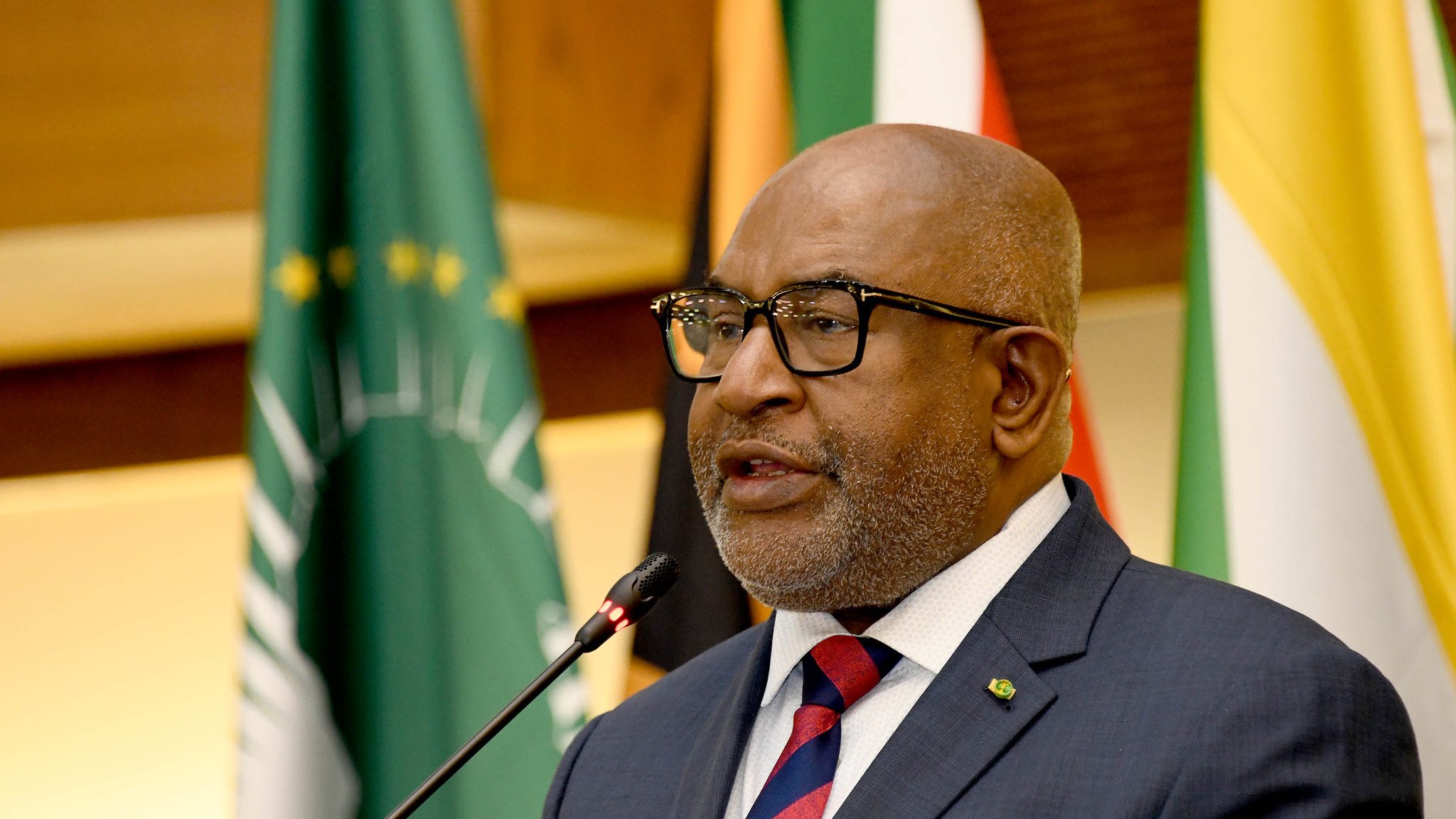 L'image représente un homme en train de prendre la parole lors d'un événement officiel. Il porte un costume sombre avec une chemise blanche et une cravate rayée, dans des tons de rouge et bleu. Ses lunettes sur le nez lui donnent une allure distinguée. Il se tient près d'un microphone, semblant s'adresser à un public. En arrière-plan, on peut apercevoir des drapeaux représentant différents pays, ce qui suggère que l'événement a une portée internationale. L'atmosphère semble solennelle et officielle, marquée par un éclairage chaleureux.