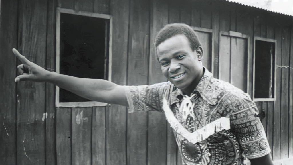 L'image montre un homme debout à côté d'une maison en bois. Il porte une chemise colorée au motif complexe, avec des nuances de marron et de beige. Son sourire est amical et engageant. L'homme pointe une main vers un mur en bois, suggérant une direction ou attirant l'attention sur quelque chose. En arrière-plan, on aperçoit des fenêtres sombres qui ajoutent une dimension de profondeur à la scène. L'atmosphère semble à la fois chaleureuse et authentique, évoquant un moment de convivialité.