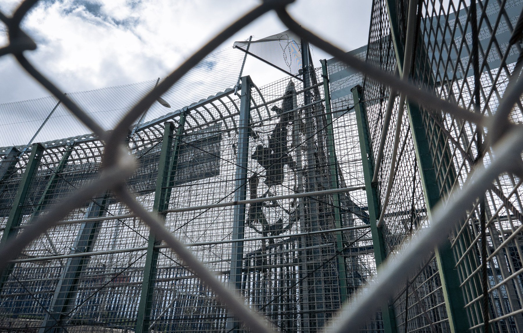 L'image représente une structure de sécurité, probablement une clôture ou un mur de protection. On aperçoit des barrières métalliques en grillage, qui sont transparentes et permettent de voir à travers. À l'intérieur, on distingue une installation, probablement en hauteur, marquée par des éléments solides tels que des poteaux. Le ciel en arrière-plan est nuageux, laissant entendre une lumière diffuse. L'ensemble dégage une impression d'isolement et de confinement, accentuée par la perspective de la clôture.